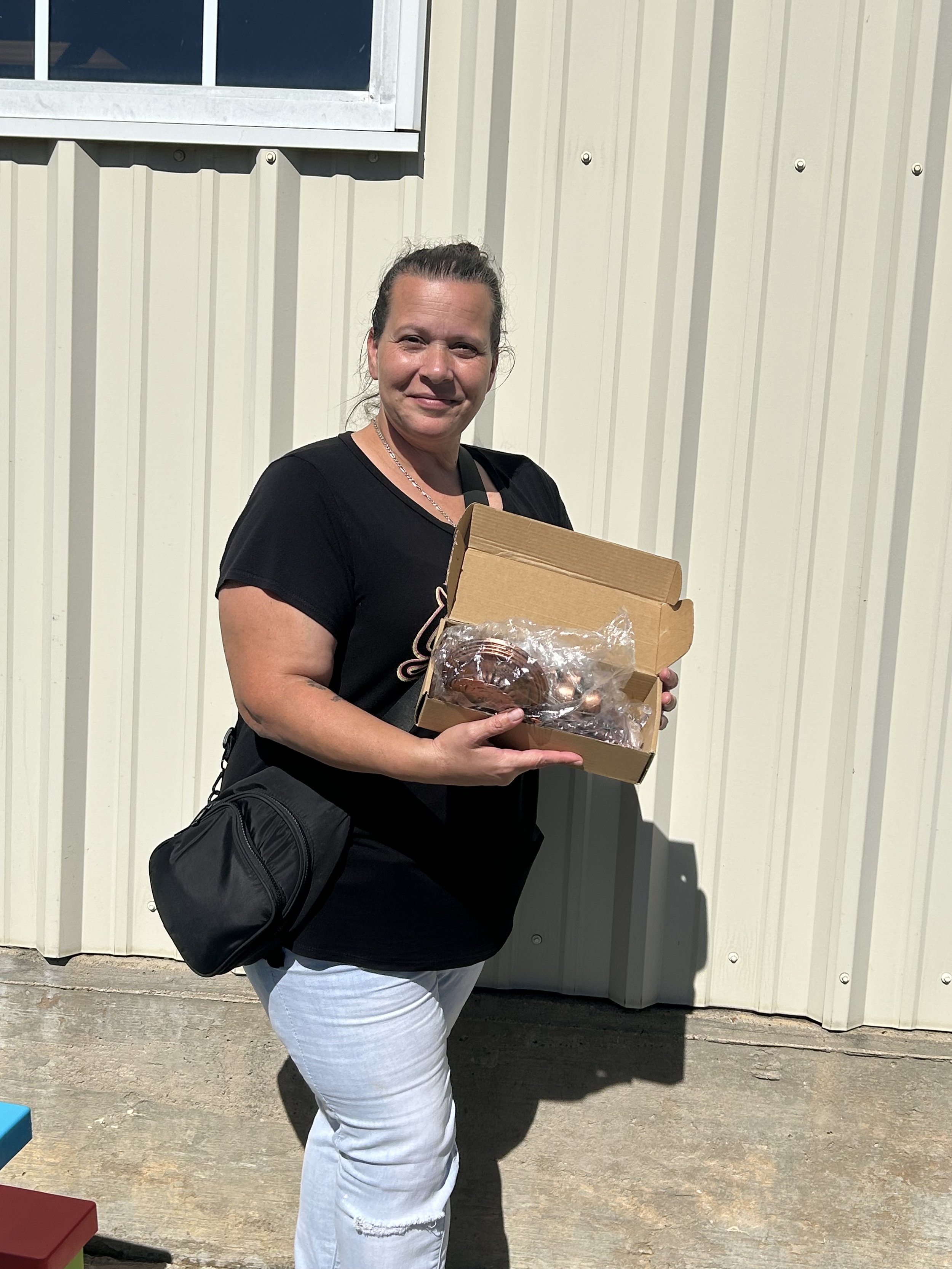 A woman holding a cardboard box with a cake and chocolates outside in front of a light-colored metal building wall.