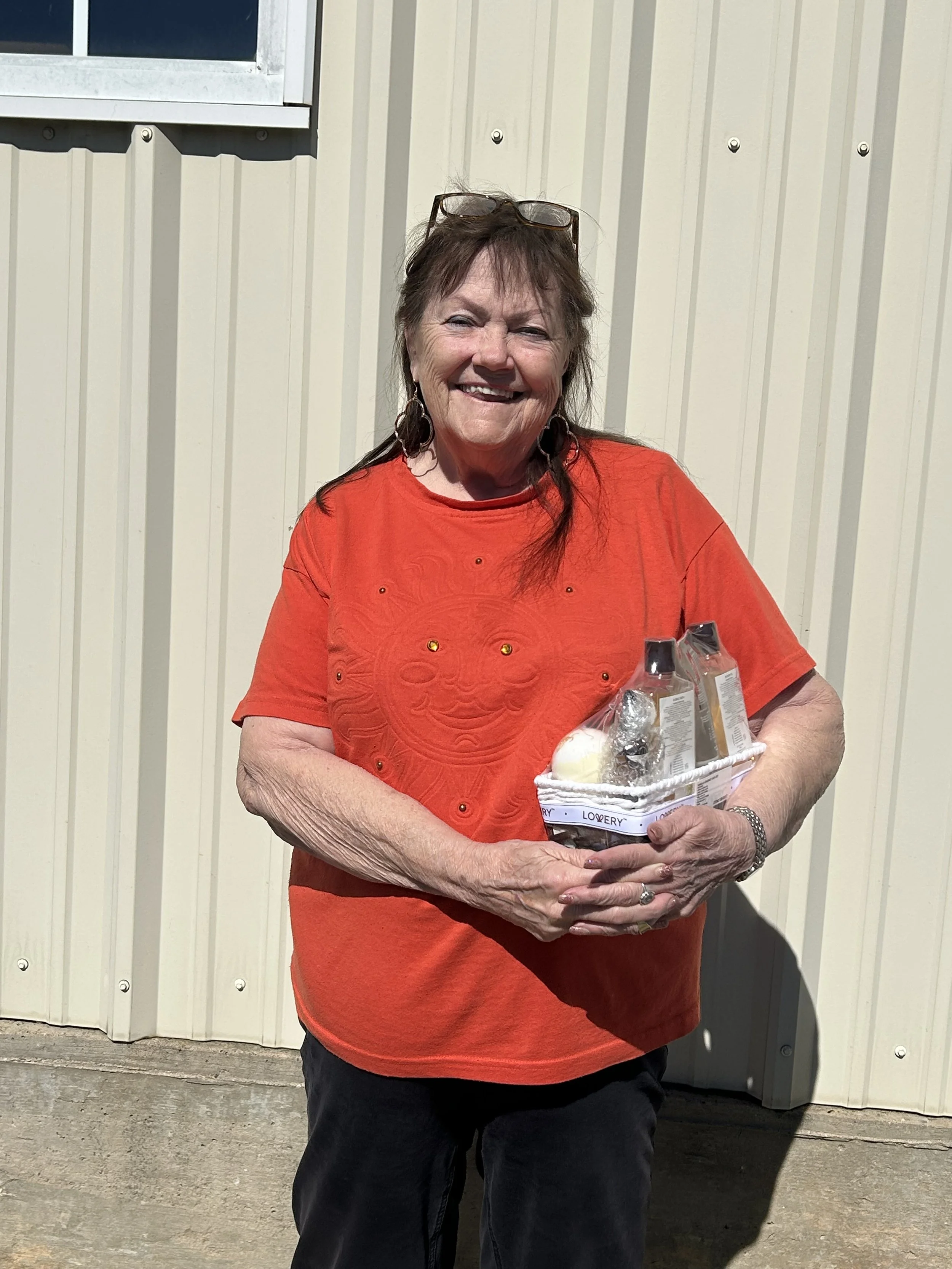 A smiling woman with glasses on her head, earrings, and wearing a red shirt holding a gift basket with bath products, standing in front of a beige metal building with a window.