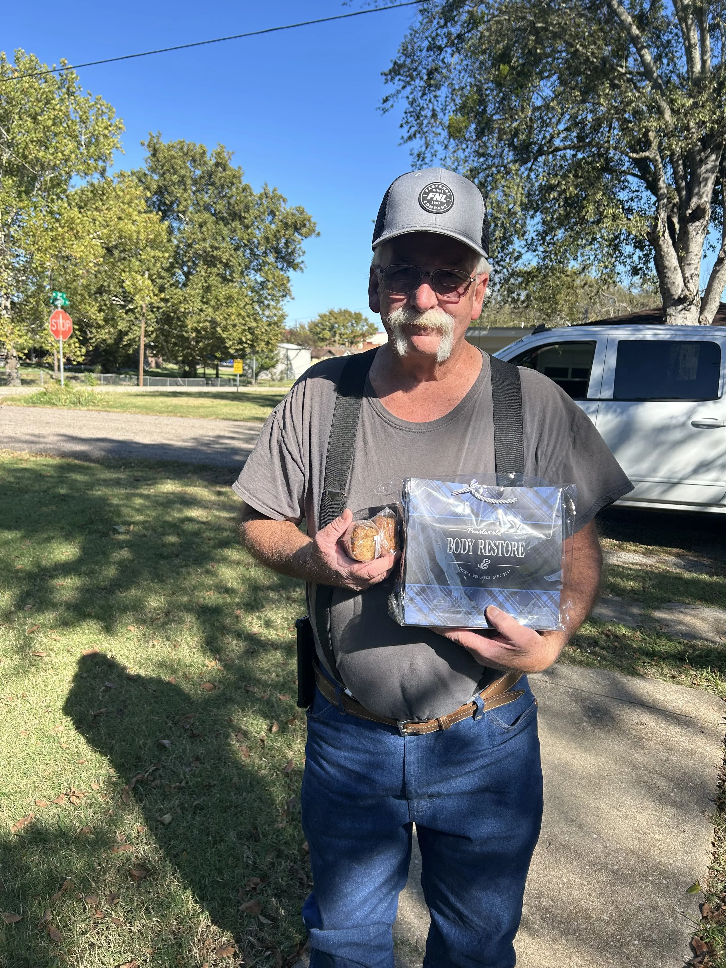 Older man with a gray baseball cap, sunglasses, and gray t-shirt holding a small packaged gift and a bag labeled 'Body Restore' outdoors on a sunny day.