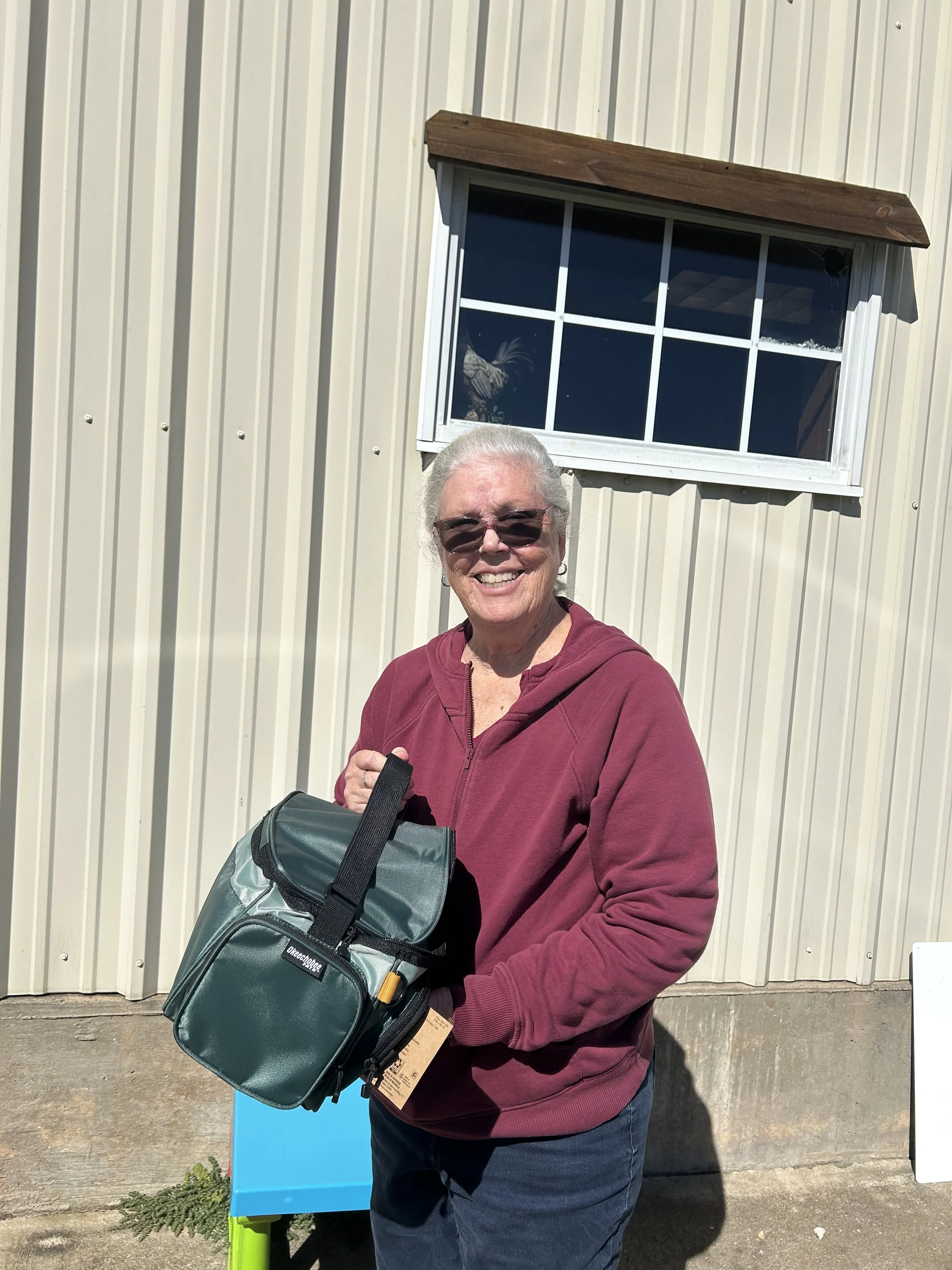 An elderly woman with gray hair wearing sunglasses, a maroon hoodie, and dark jeans, smiling and holding a green insulated lunch bag, standing outside in front of a cream-colored metal building with a window and wooden awning.