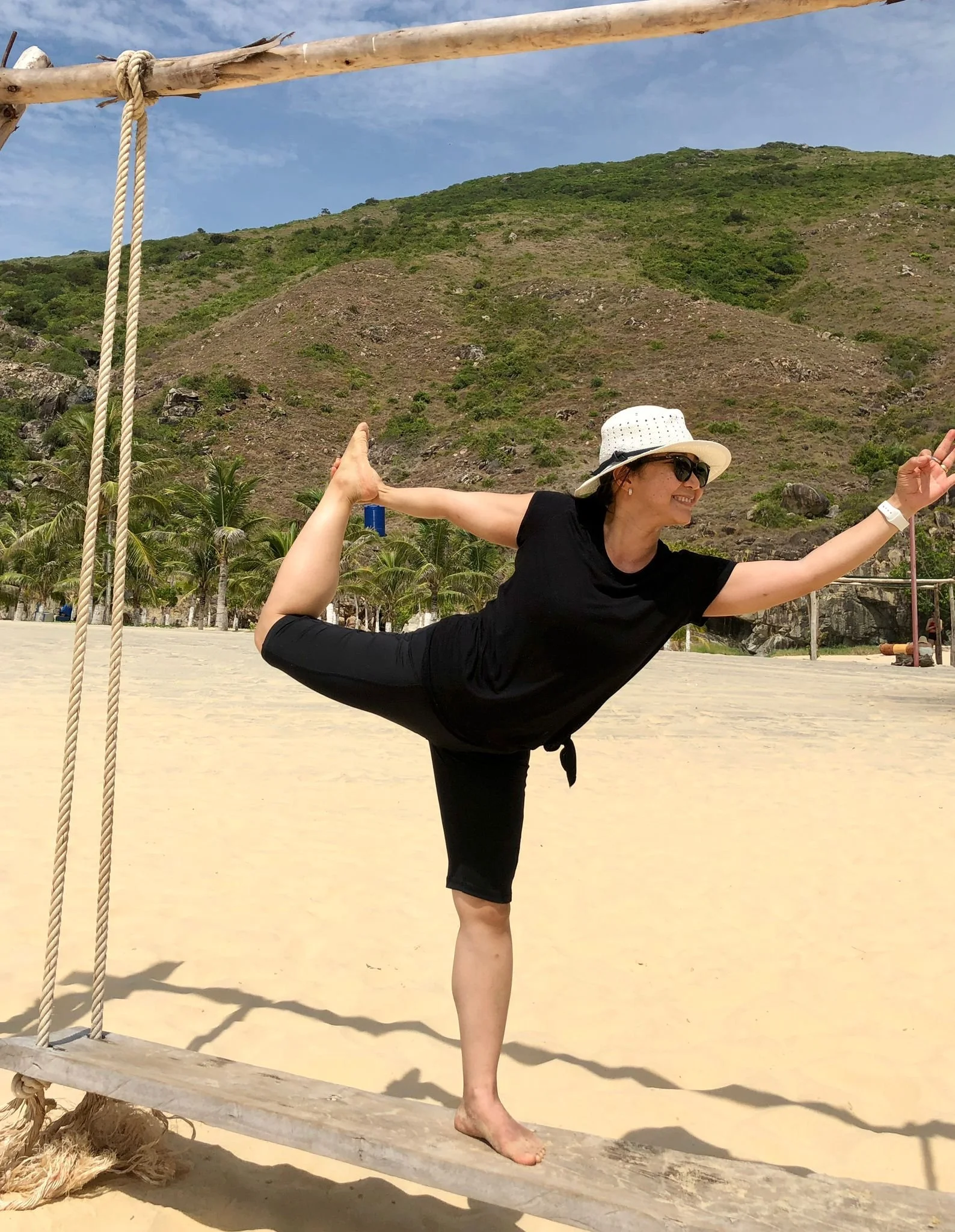 Une femme faisant du yoga sur un pont en bois suspendu à la plage, portant un chapeau blanc et des lunettes de soleil, avec un arrière-plan de collines et de palmiers.