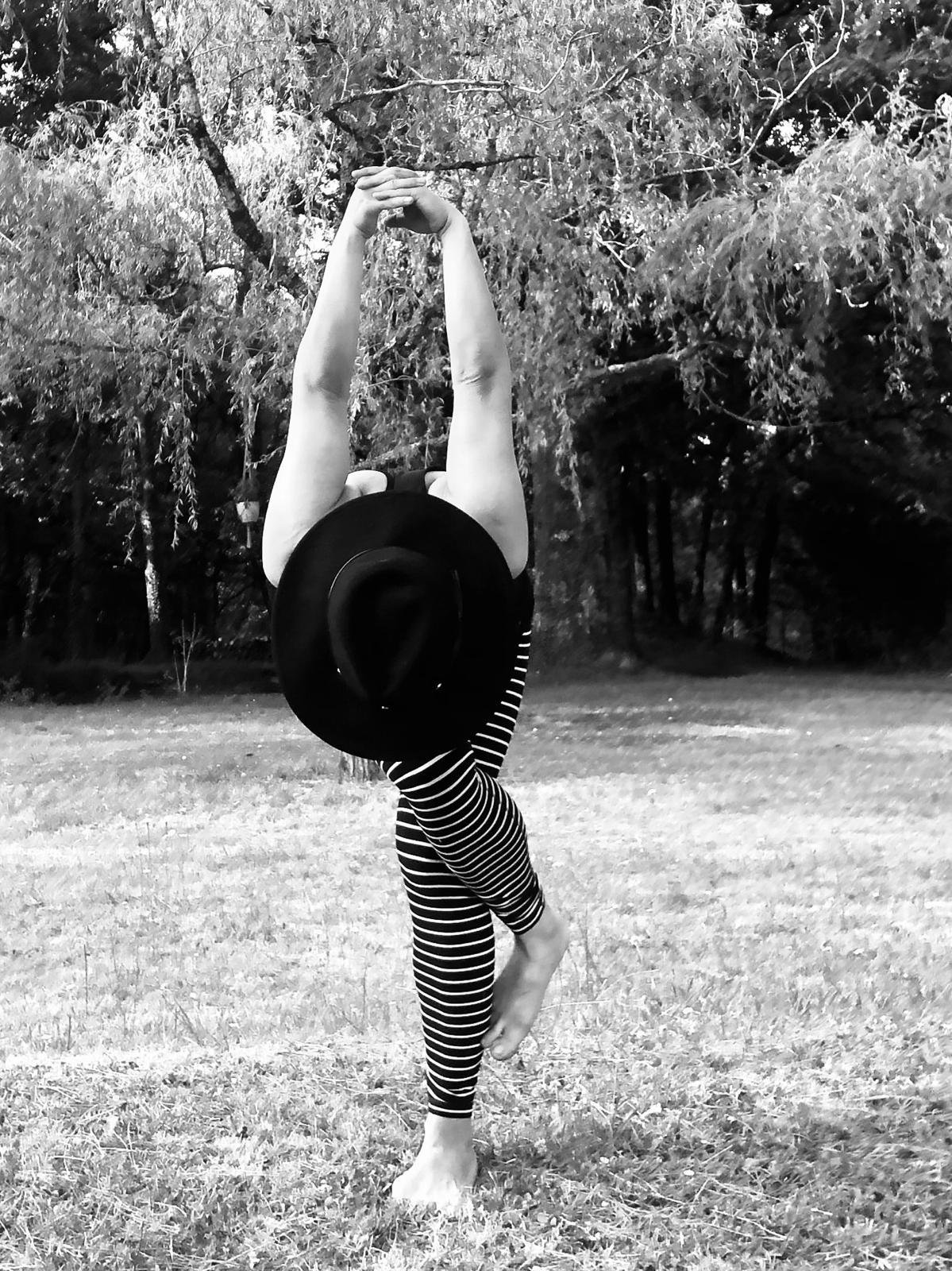 Une personne effectuant une pose de yoga ou d'exercice en équilibre sur une jambe dans un parc, portant un chapeau noir et un pantalon rayé, avec des arbres en arrière-plan.