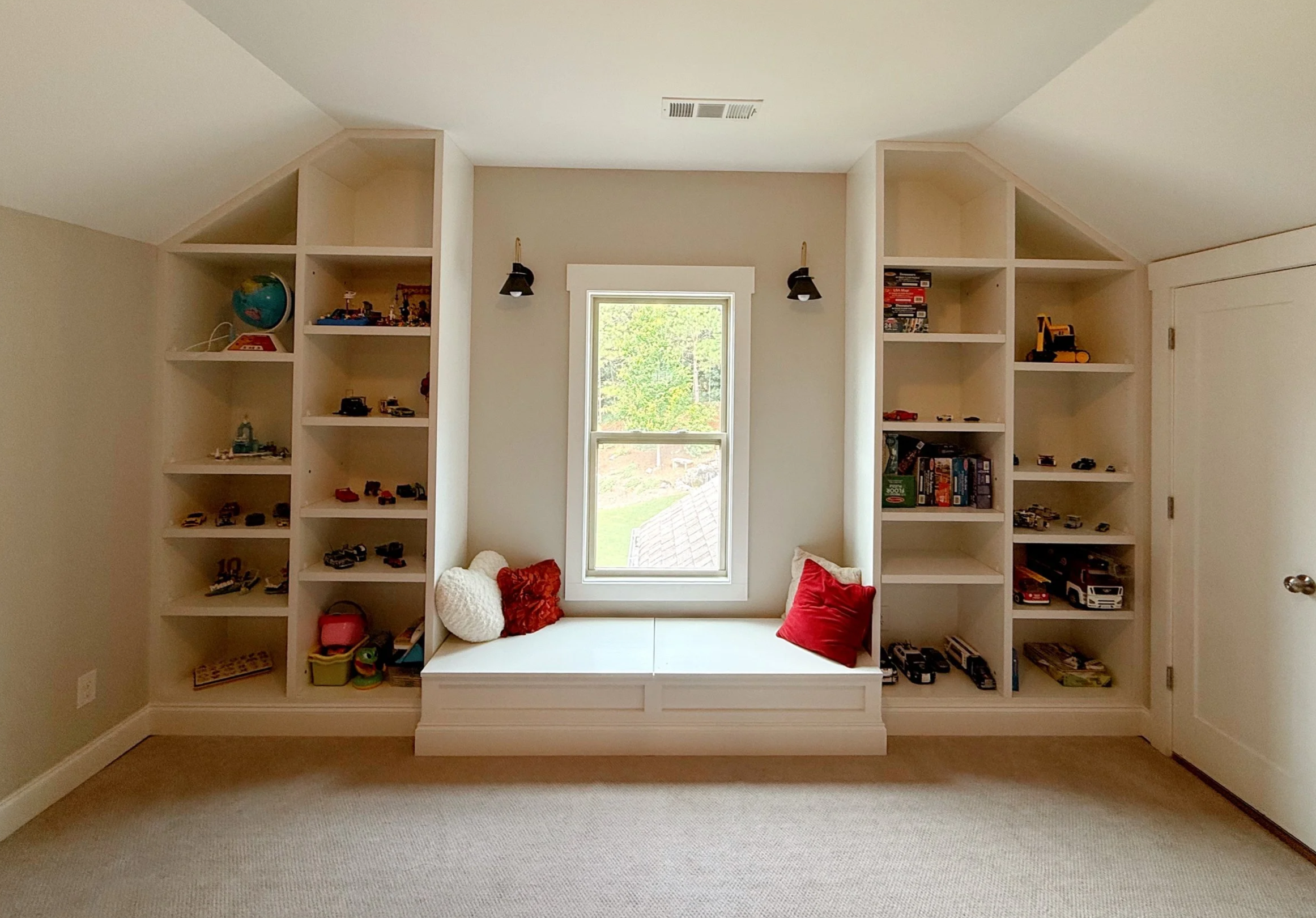 A built-in white bookshelf with an arched seating area in the center, adorned with red and white cushions. The shelves contain toys, books, and decorative items, and there is a window with a view of greenery behind it. Two black wall sconces flank the window.