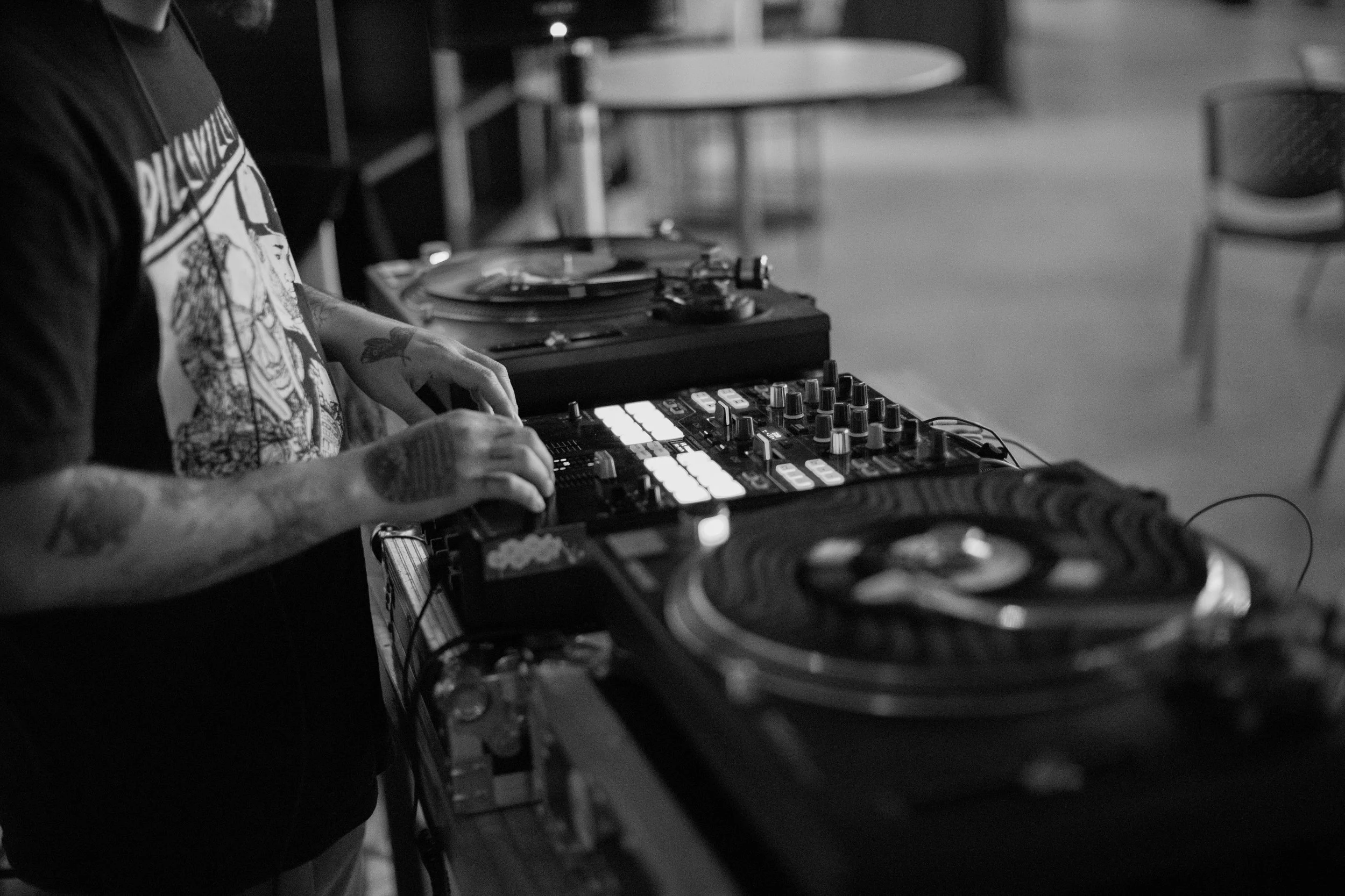 A person with tattoos is DJing with a turntable and mixer, in a black and white photo.