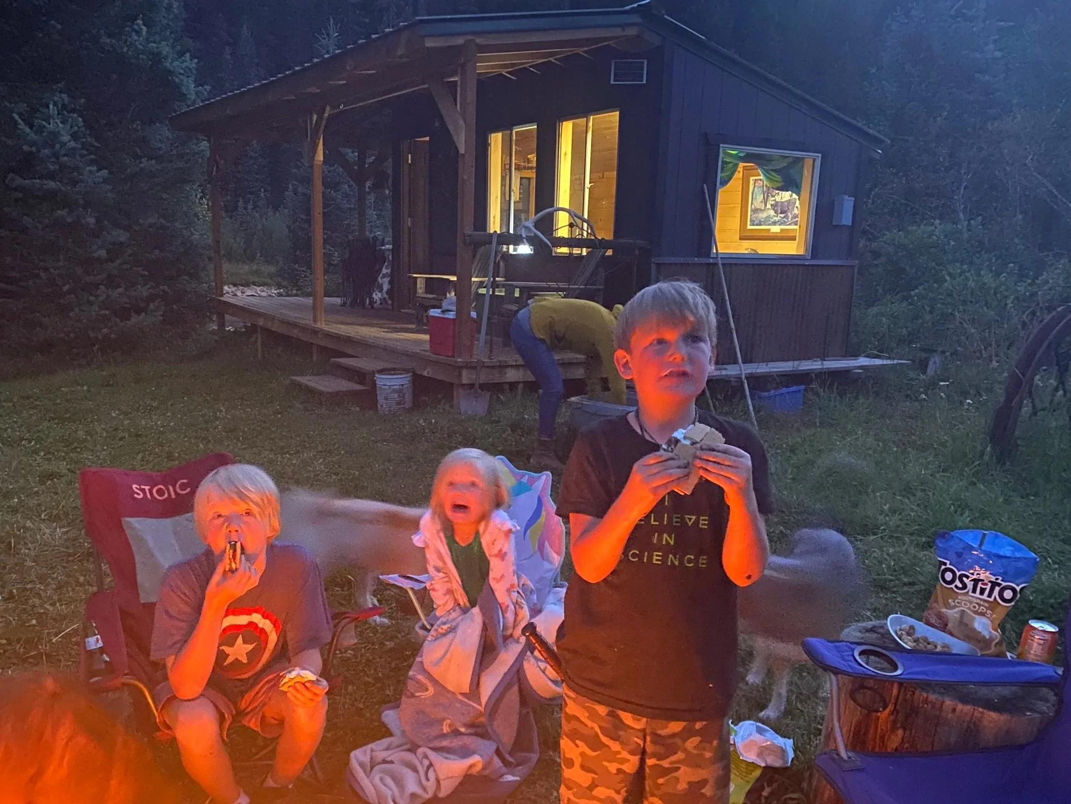 Children sitting outside near a wood cabin during evening, some eating snacks, with a woman in the background bending over, and a cow on the porch.
