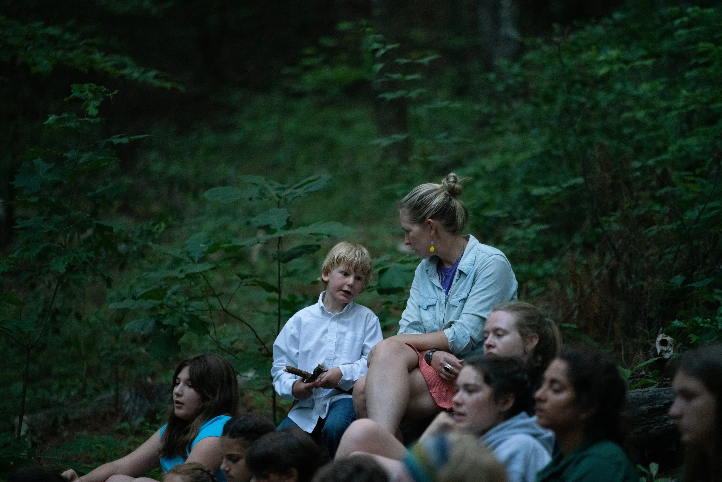 A group of children and a woman sitting on a log in a dense, green forest, with trees and foliage surrounding them.