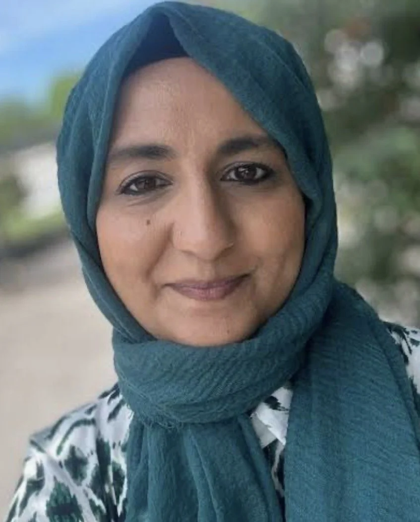 Close-up of a woman wearing a teal headscarf, smiling outdoors with blurred trees in the background.