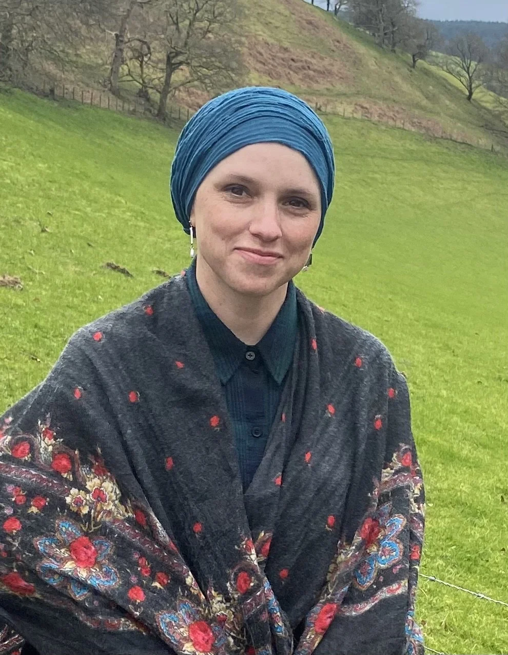 A woman with fair skin and brown eyes wearing a teal headwrap, earrings, a dark button-up shirt, and an ornate black shawl with red, blue, and gold floral patterns standing outdoors on a grassy hill with trees and a fence in the background.