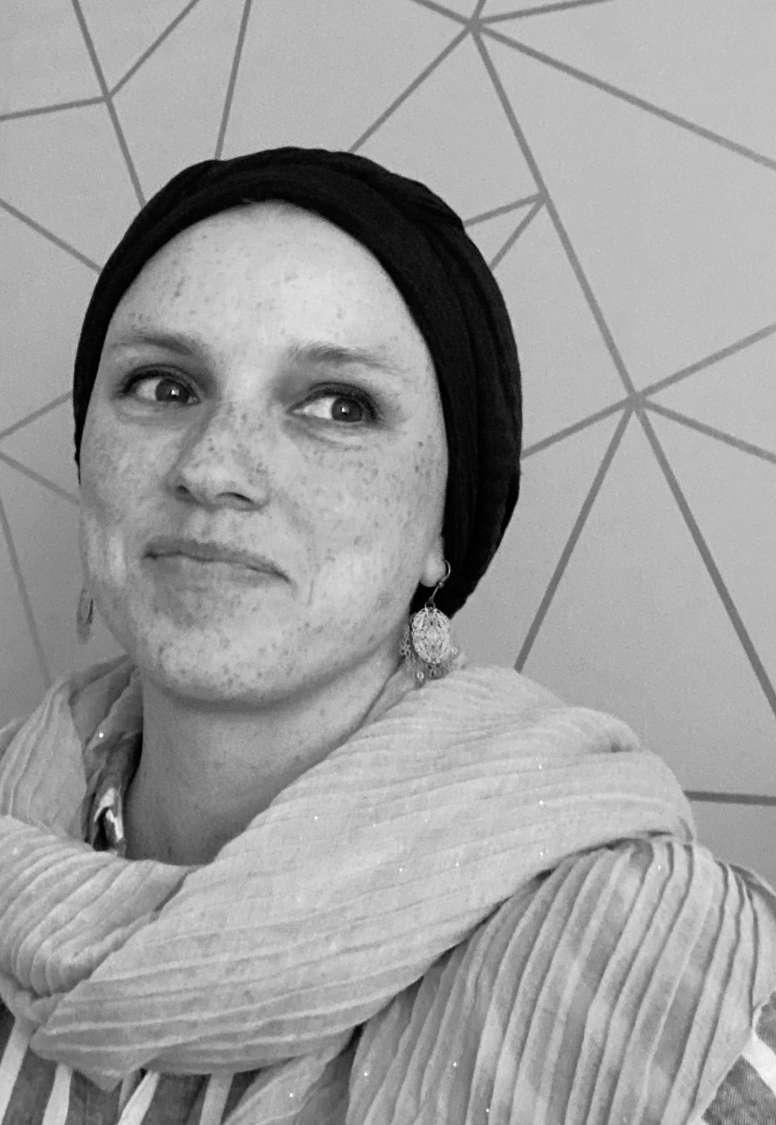 A woman with freckles wearing a headscarf and earrings, looking to the side, with a geometric pattern on the background ceiling.