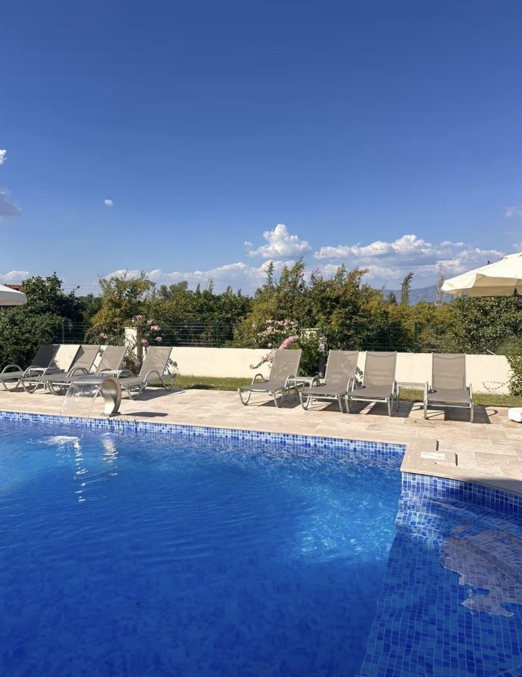 Swimming pool with blue water, lounge chairs, white umbrellas, greenery, and a clear blue sky with some clouds.