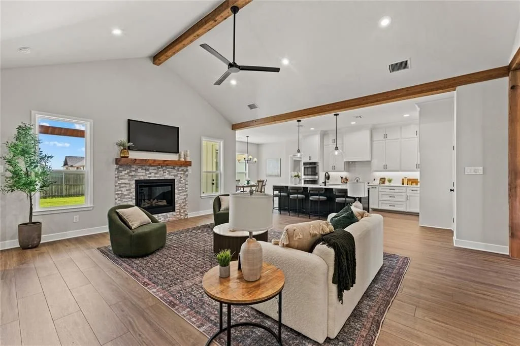 Open-concept living room and kitchen with vaulted ceiling, white walls, wooden beams, hardwood floors, fireplace, television, sofa, armchair, and kitchen island with bar stools.