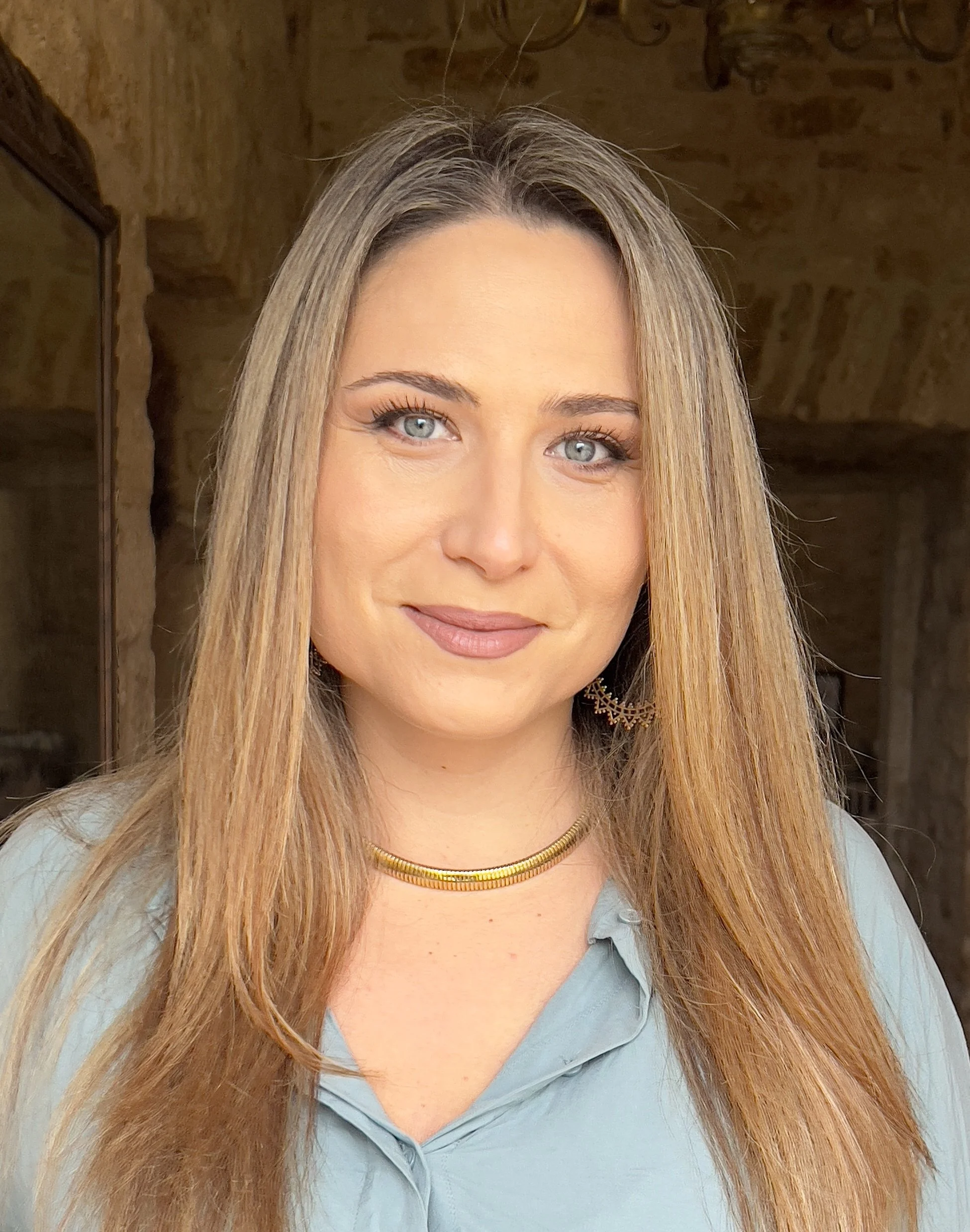 A woman with long, straight blonde hair and blue eyes smiling, wearing a light blue blouse, gold necklace, and earrings, in a rustic indoor setting with stone walls.