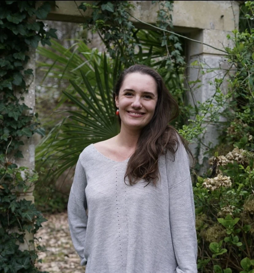 A woman with long brown hair and red earrings smiling outdoors among green plants and stone pillars.