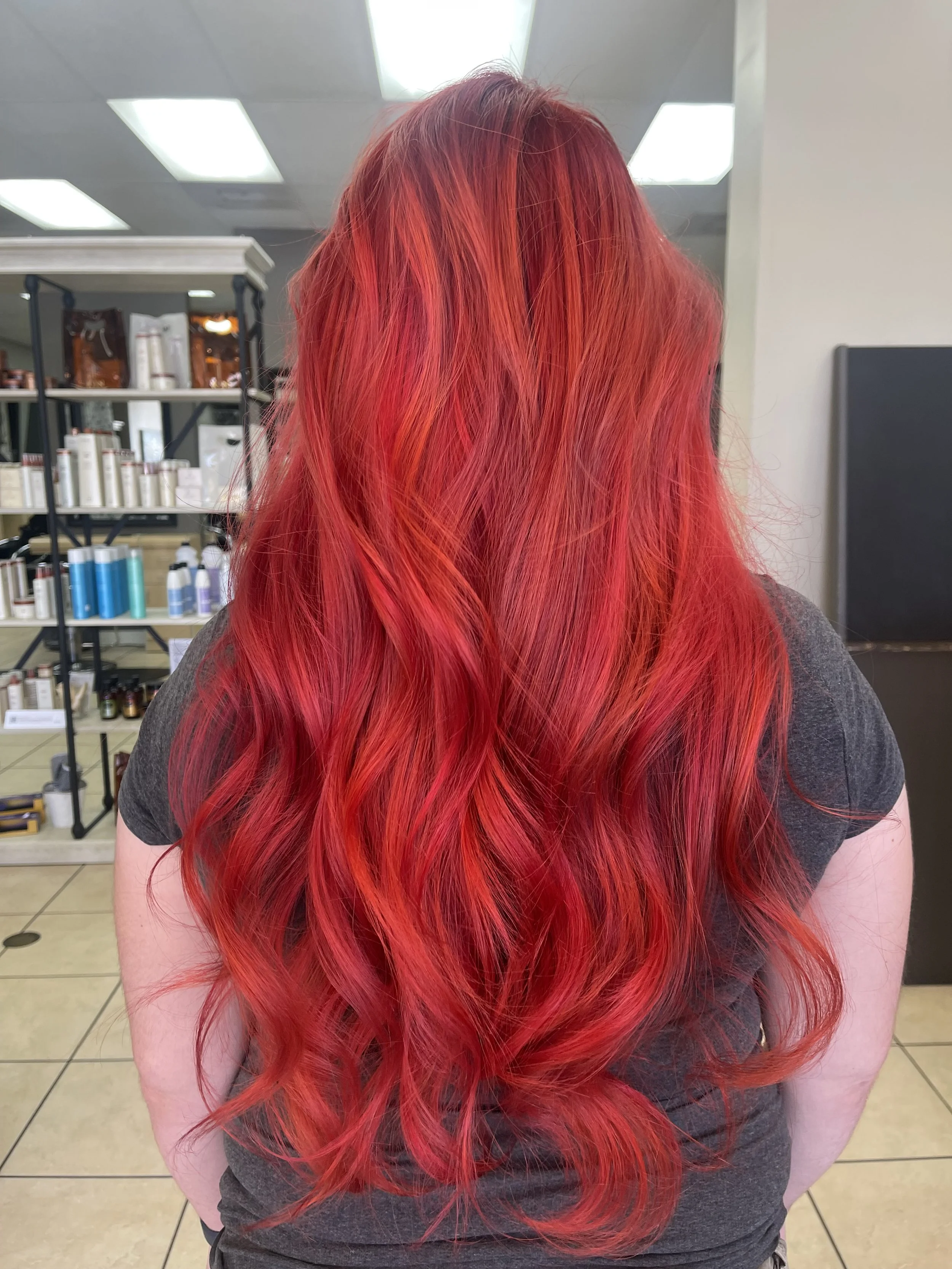 Back view of a woman with long, wavy, bright red hair, standing in a salon with shelves of hair products in the background. hair colorist Martin Rodriguez
