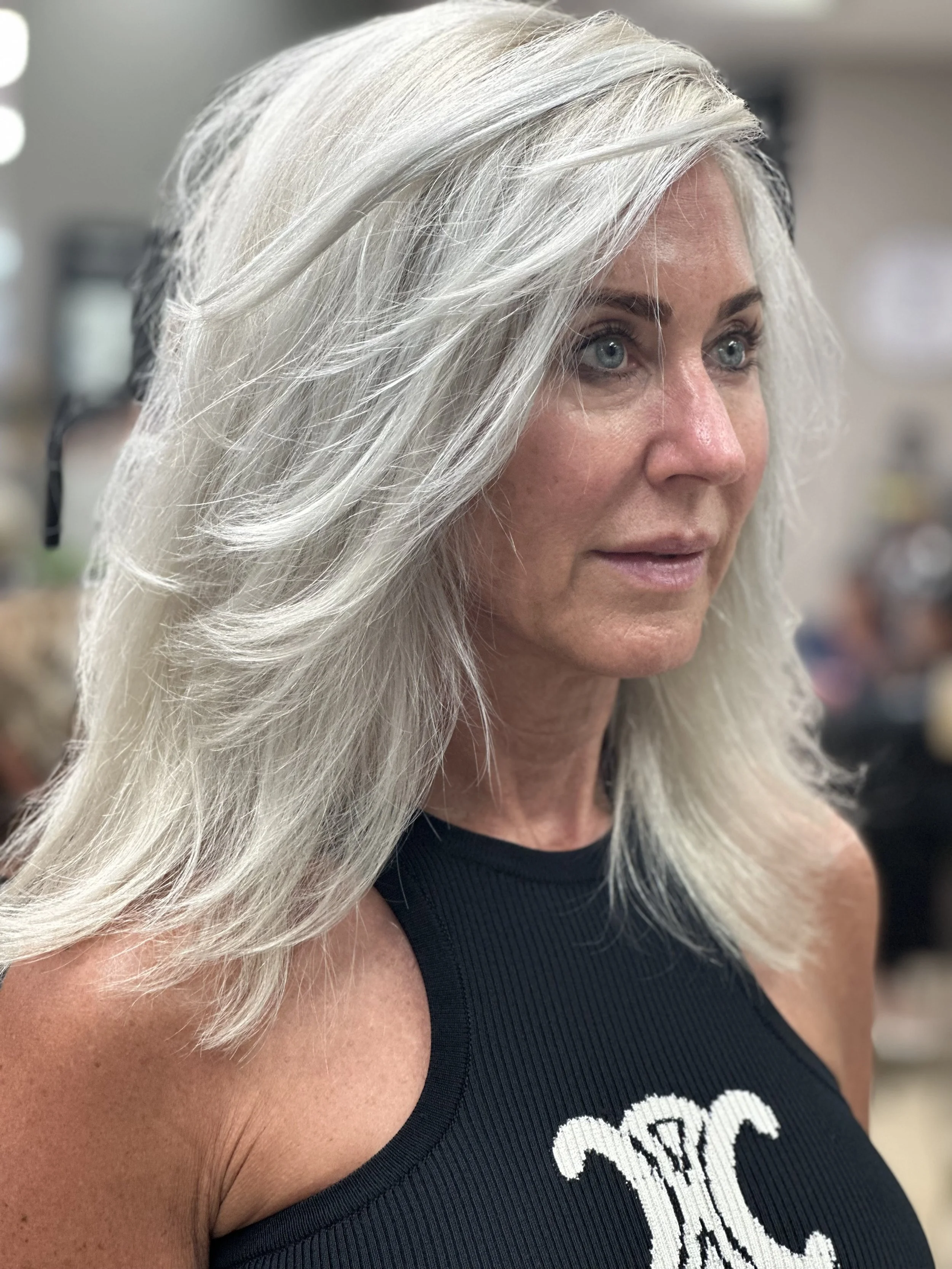 A woman with long, wavy gray hair and blue eyes wearing a black top with white embroidery, standing indoors with a blurred background.