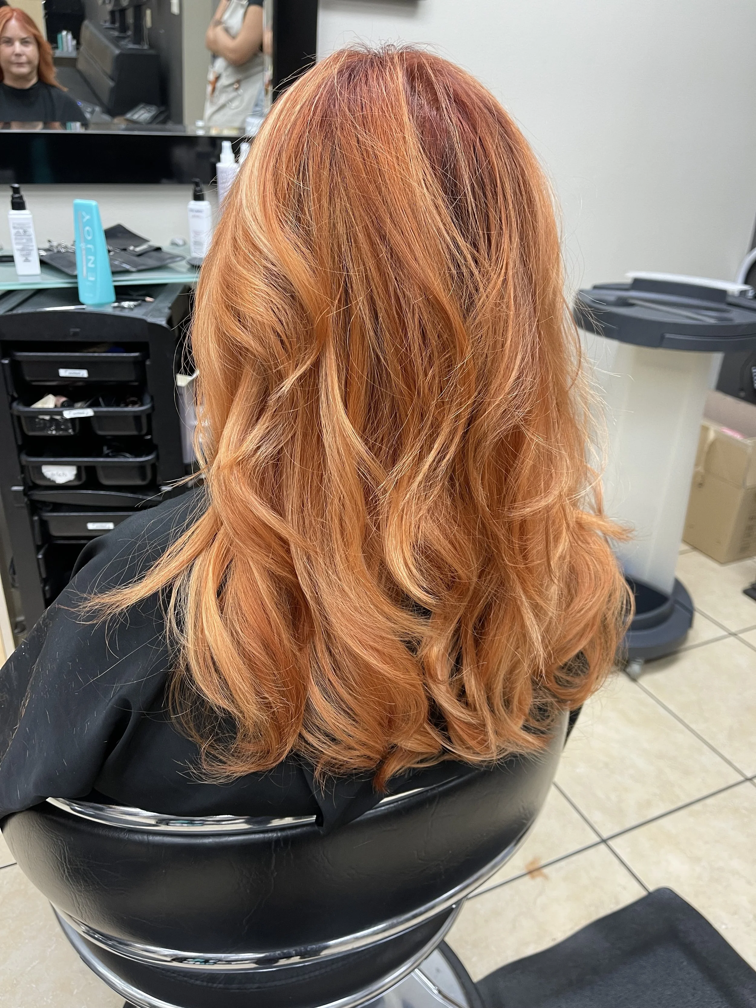 Back view of a woman with long, wavy, reddish-orange hair sitting in a hair salon chair.