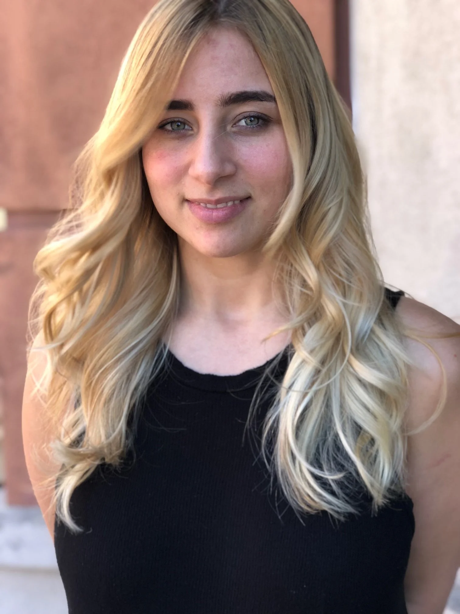 A young woman with blonde wavy hair and blue eyes smiling while wearing a black sleeveless top, standing outdoors with a blurred background.