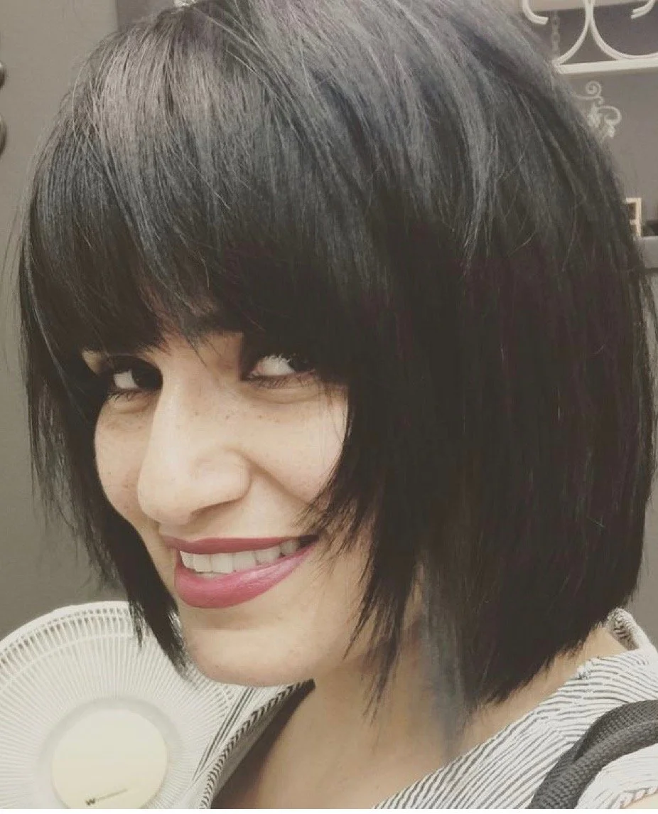 A woman with short black hair smiling, with her head tilted slightly to the right, in front of a white fan.