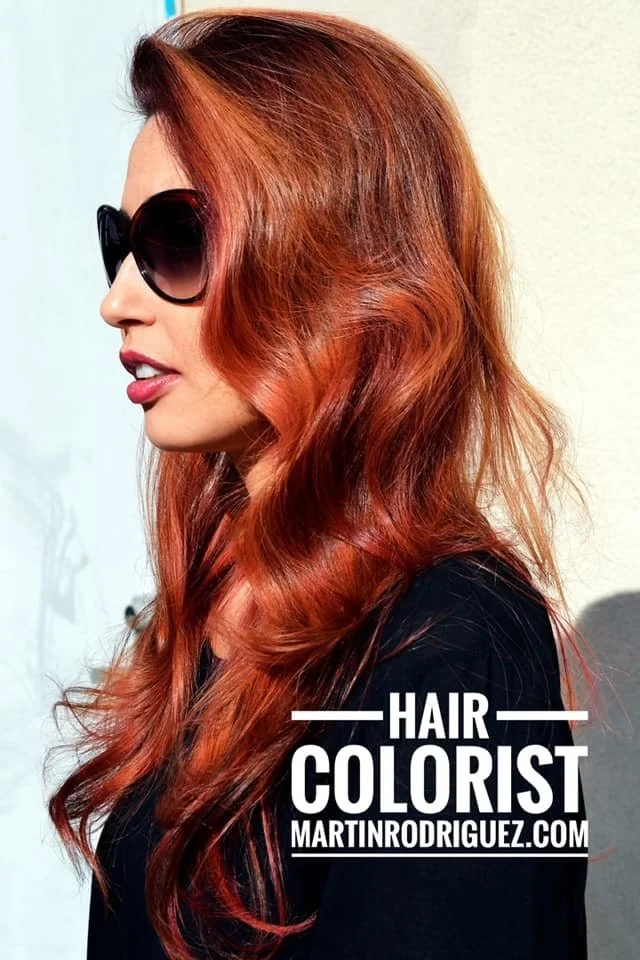 A woman with long wavy red hair and wearing large black sunglasses, looking to the left, standing outside against a light background, with the text "HAIR COLORIST MARTINRODRIGUEZ.COM" at the bottom.