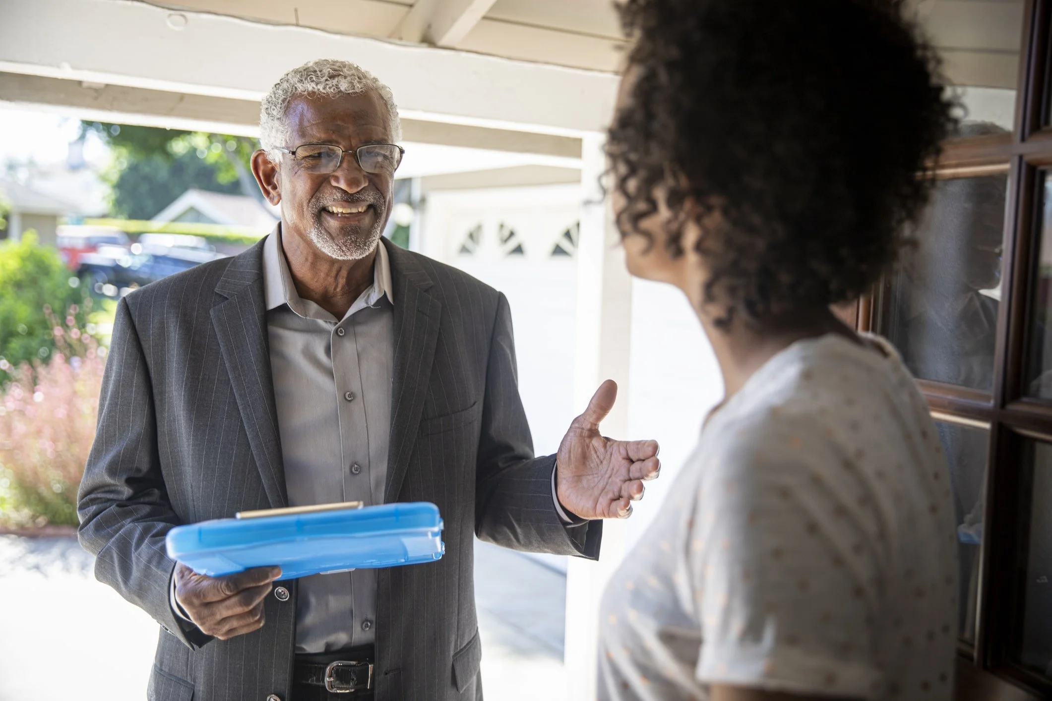 An elderly man in a gray suit and glasses talking to a woman, holding a small blue box, outside in a residential area.