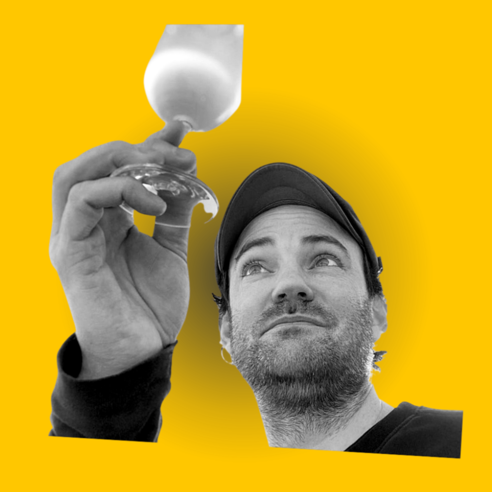 A man with a beard and cap holding a wine glass against a yellow background.