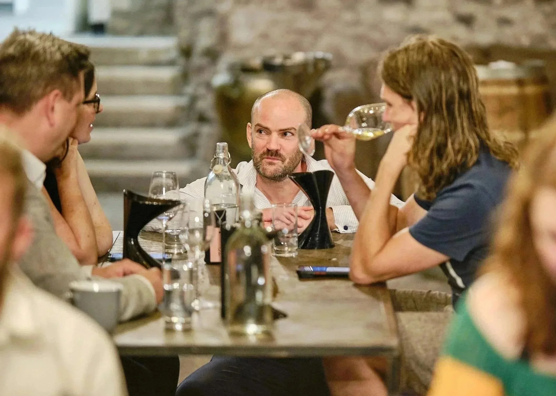 A group of people at a dinner table engaged in conversation, with one woman drinking wine and a man looking annoyed or suspicious.