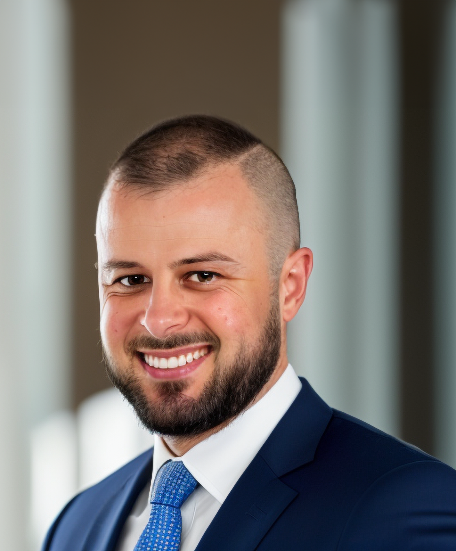 A portrait of a smiling man with a neat beard and short hair, wearing a dark blue suit, white shirt, and blue tie, with a blurred background of pillars or columns.