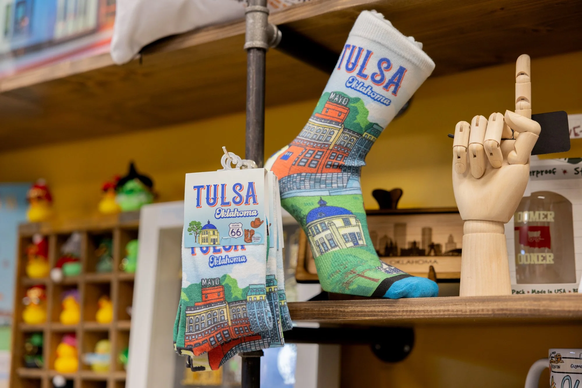 A sock decorated with colorful illustrations of Tulsa, Oklahoma landmarks, displayed on a stand next to a wooden mannequin hand making a 'rock on' gesture.