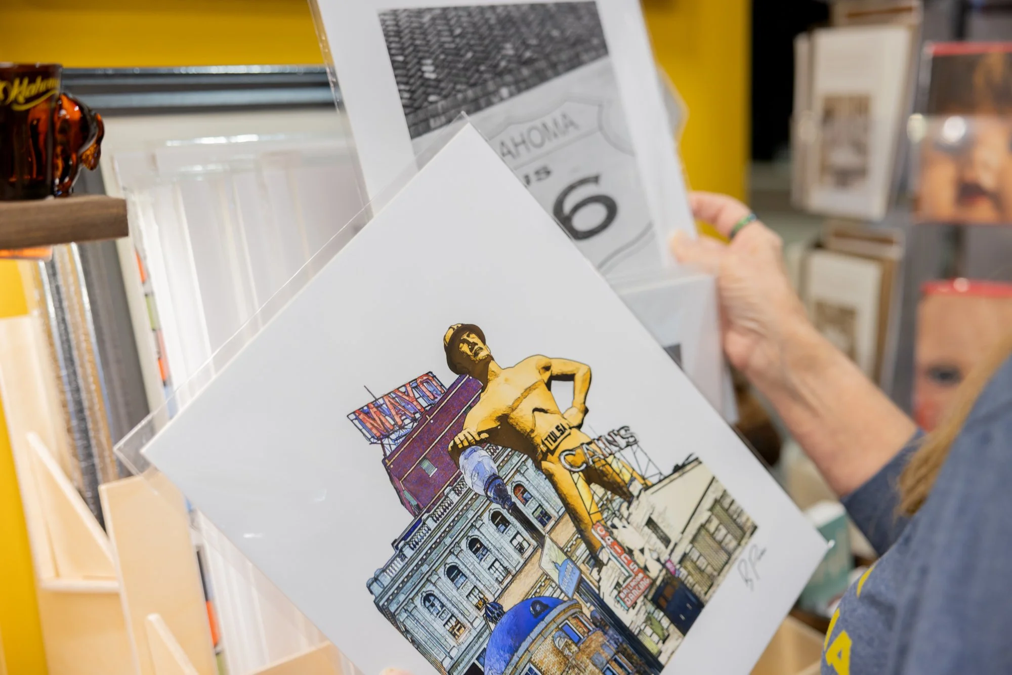 Person browsing postcards at a store, with a card featuring a drawing of a topless man standing in front of a city building, holding a skateboard, against a backdrop of city buildings and signs.