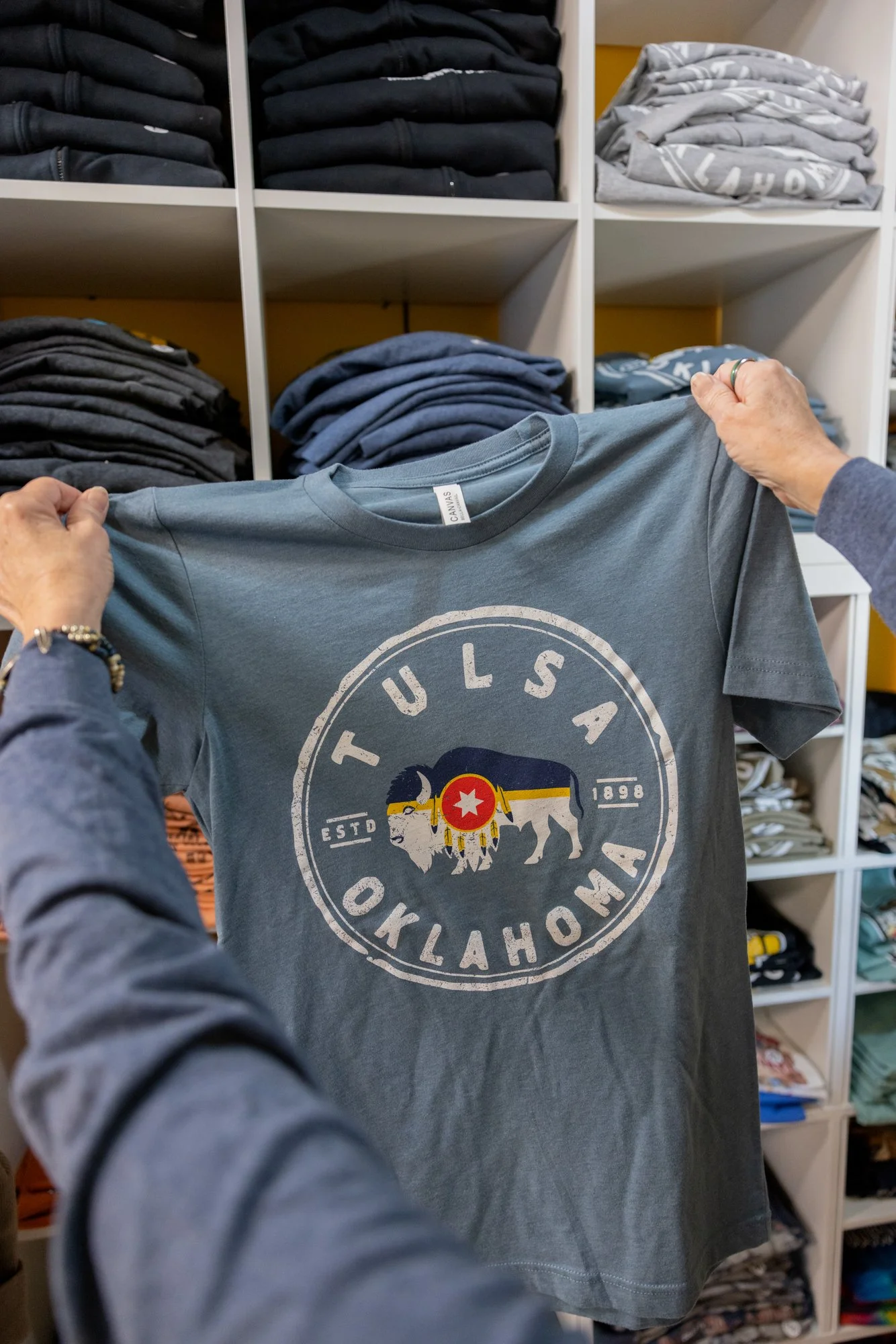 A person holding a gray T-shirt with the Tulsa, Oklahoma logo in front of shelves filled with folded clothes.