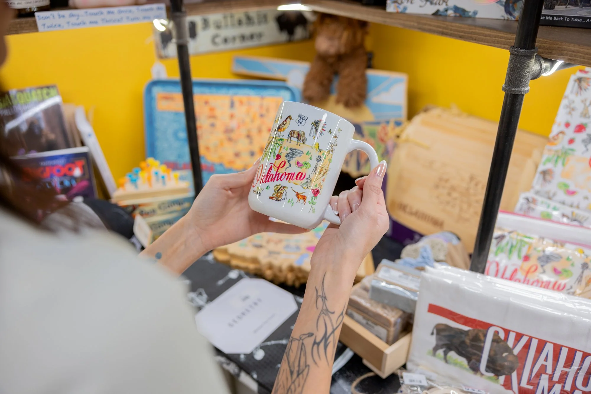 A woman holding a white ceramic mug with colorful illustrations and the word 'Oklahoma' on it, at a souvenir shop with various other merchandise and books on shelves in the background.