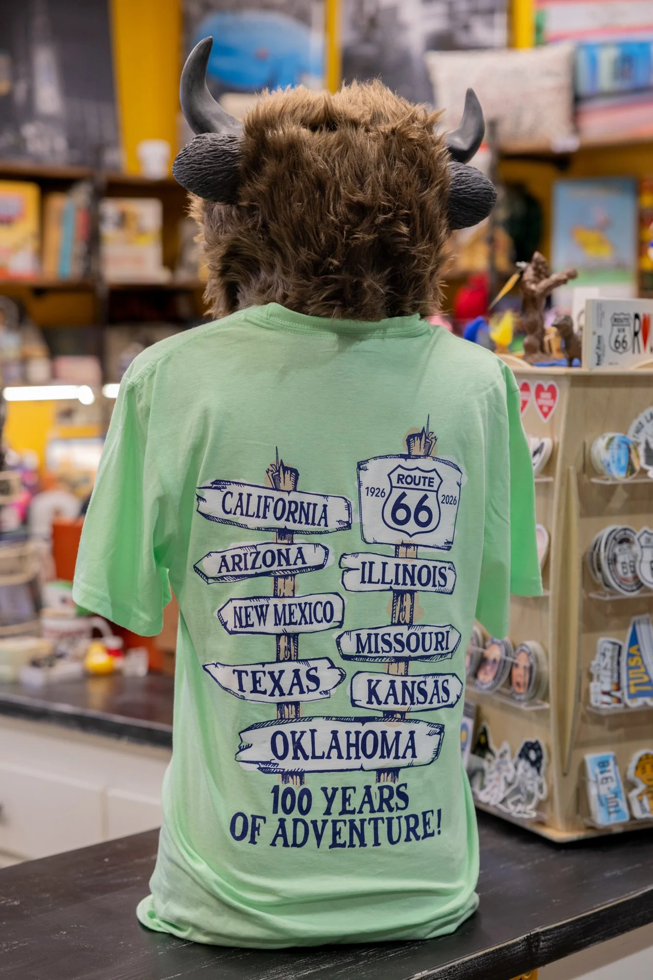 Person wearing a costume with a buffalo head and horns, dressed in a green T-shirt with signposts pointing to various states and Route 66, at a store with shelves of souvenirs and trinkets.