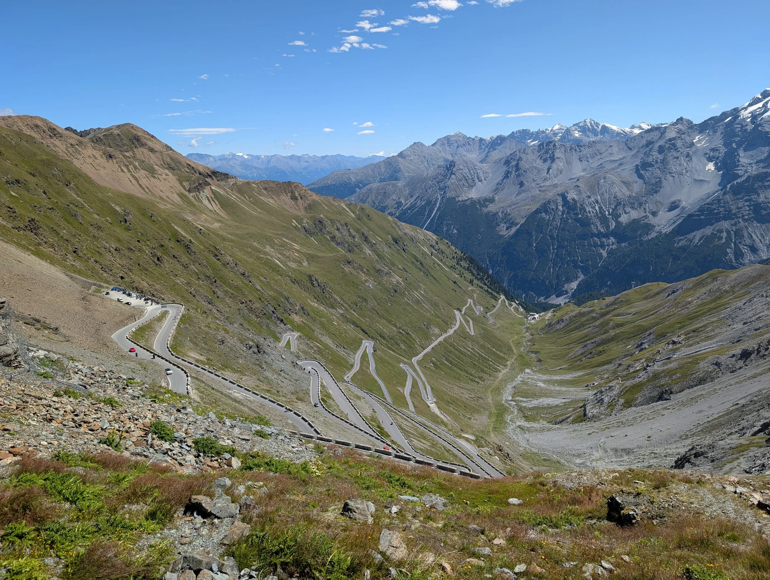 Berglandschaft mit Serpentinenstraße, grünem Gras, felsigen Hügeln und hohen Bergen im Hintergrund unter blauem Himmel.