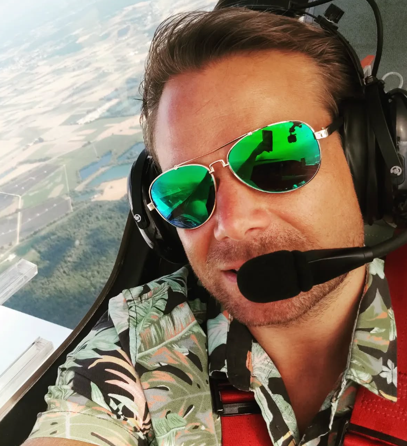 Pilot trägt dunkle Sonnenbrille mit grünen Spiegelgläsern und Headset, fliegt in einem Flugzeug über grünen Landschaften.