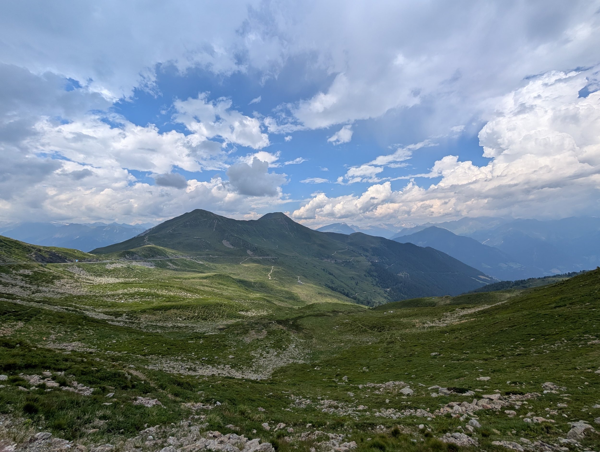 Berglandschaft mit grünen Hügeln, Bergen im Hintergrund und einem bewölkten Himmel.