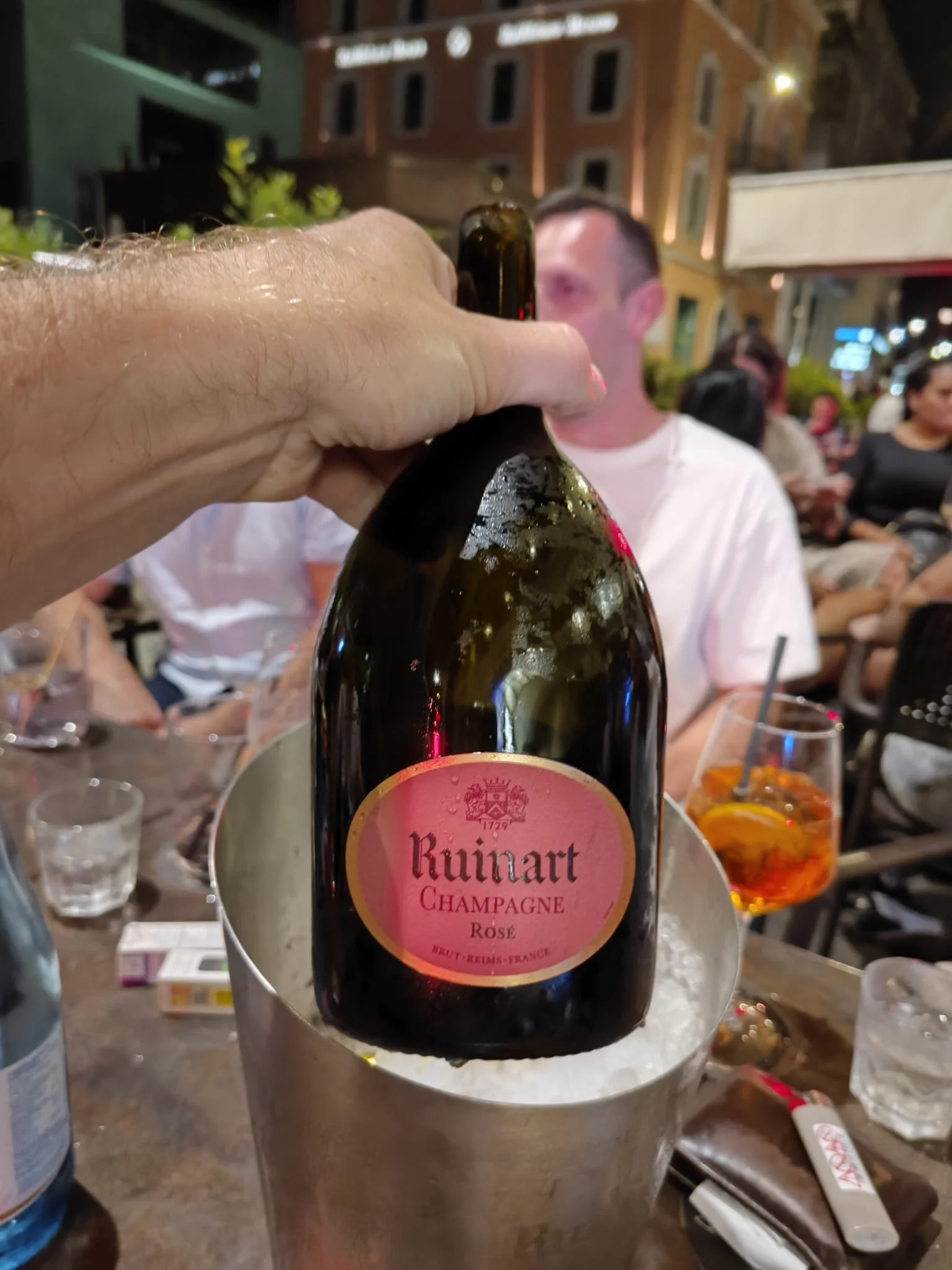 Hand hält eine Flasche Ruinart Champagner Rosé über einen Eisbehälter auf einer Party im Freien, mit Menschen im Hintergrund, Abendlicht und Gebäude.