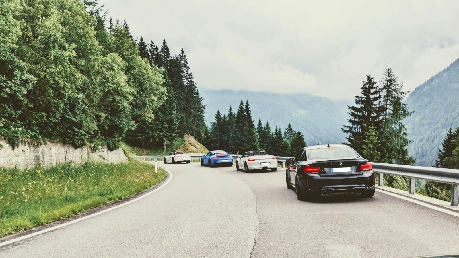 Kurvige Bergstraße mit mehreren Autos, umgeben von grünen Tannen und Bergen im Hintergrund.