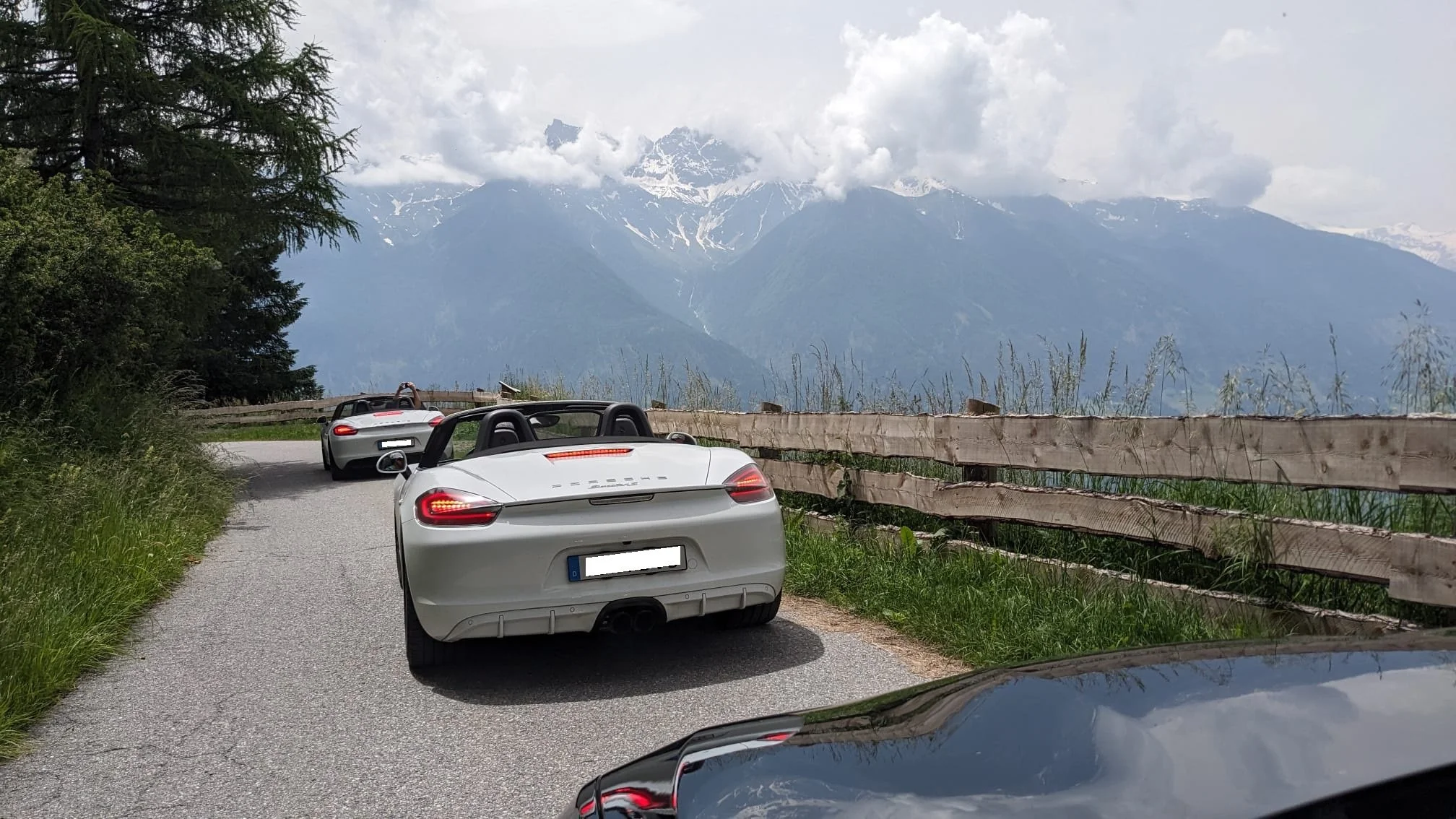 Zwei weiße Porsche-Sportwagen fahren auf einer kurvigen Bergstraße mit Holzgeländer, umgeben von grünen Bäumen und hohen Bergen im Hintergrund, teilweise schneebedeckt, bei bewölktem Himmel.