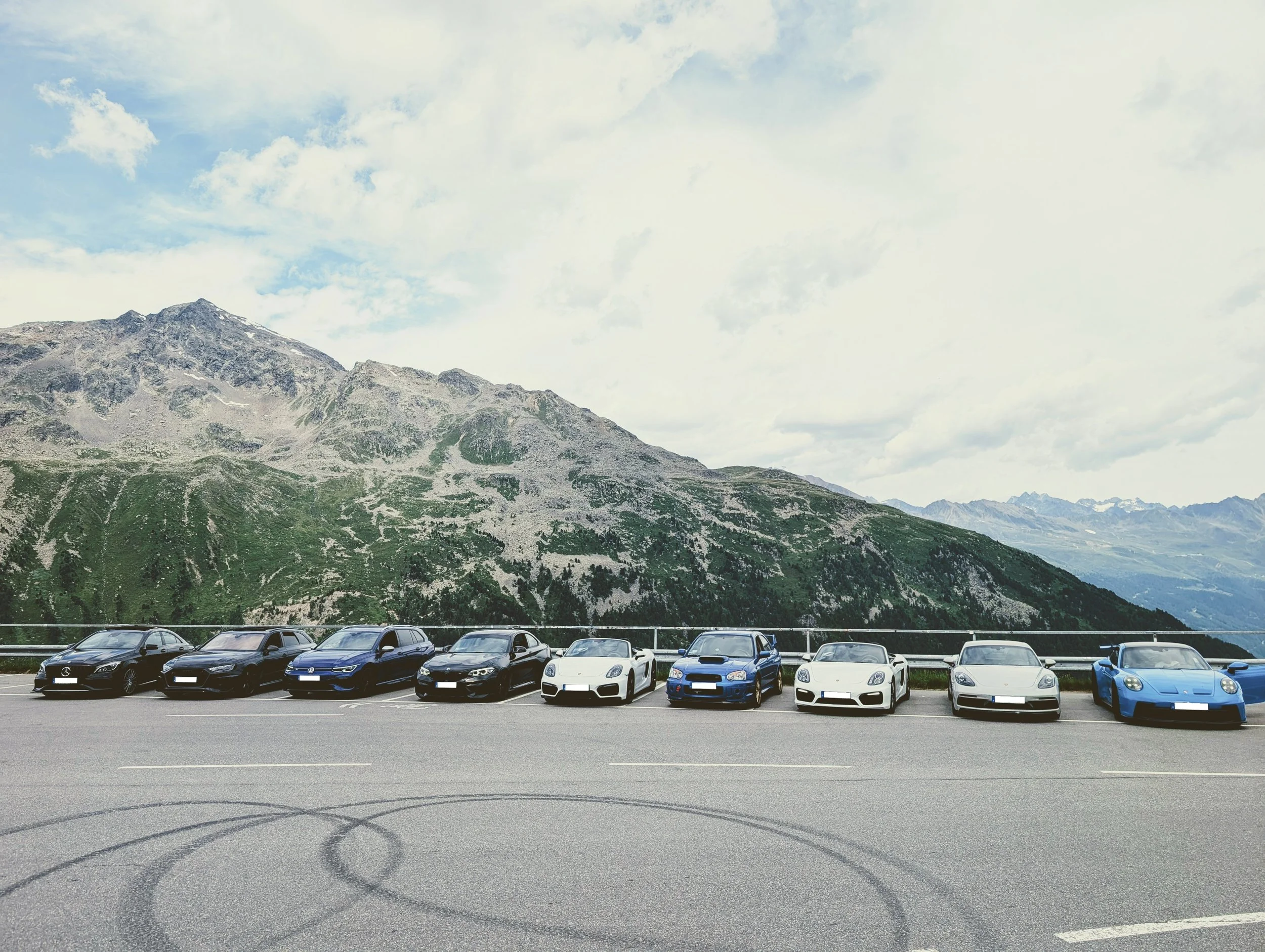 Mehrere Sportwagen und Fahrzeuge parken in einer Berglandschaft mit Wolken am Himmel.