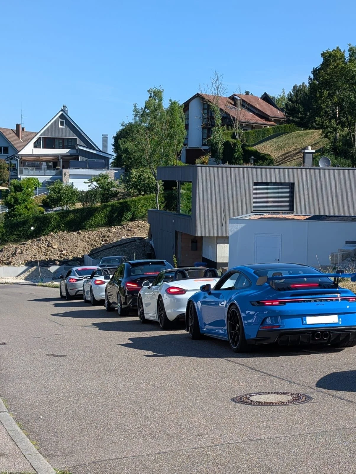 Mehrere Sportwagen, darunter ein blauer Porsche, sind auf einer Straße in einer gehobenen Wohngegend geparkt.