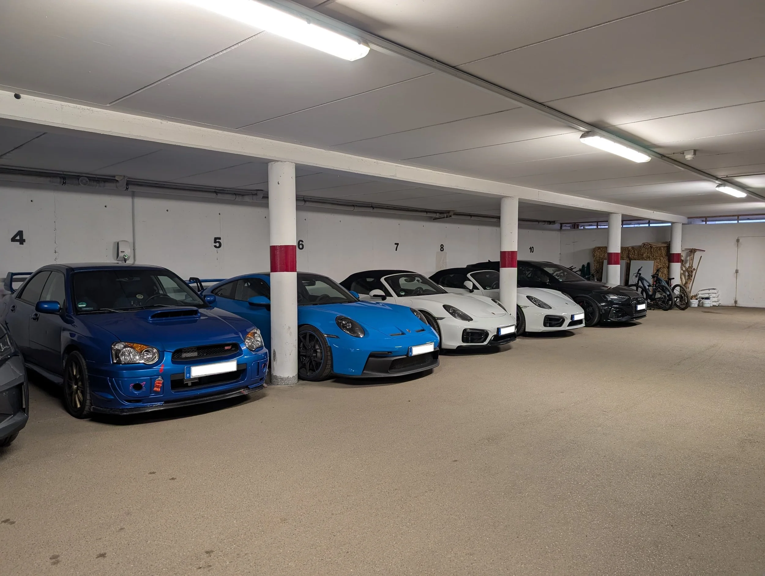 Parkhaus mit sieben Autos, darunter zwei blaue und drei weiße Sportwagen, sowie Fahrrädern und einigen Gegenständen am Ende der Reihe.