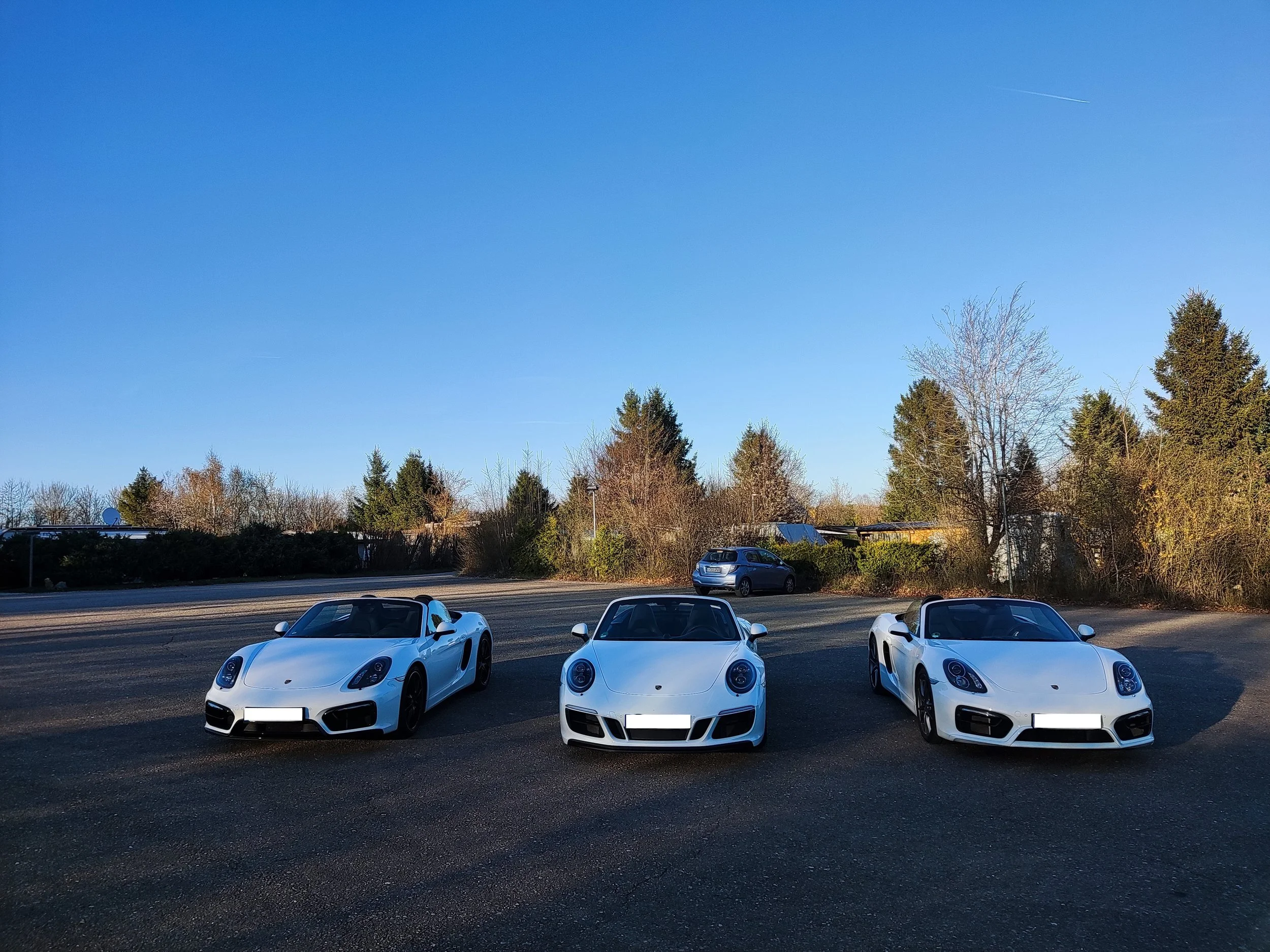 Drei weiße Porsche-Sportwagen parkend auf einem asphaltierten Parkplatz bei sonnigem Wetter, umgeben von Bäumen und einem blauen Himmel.