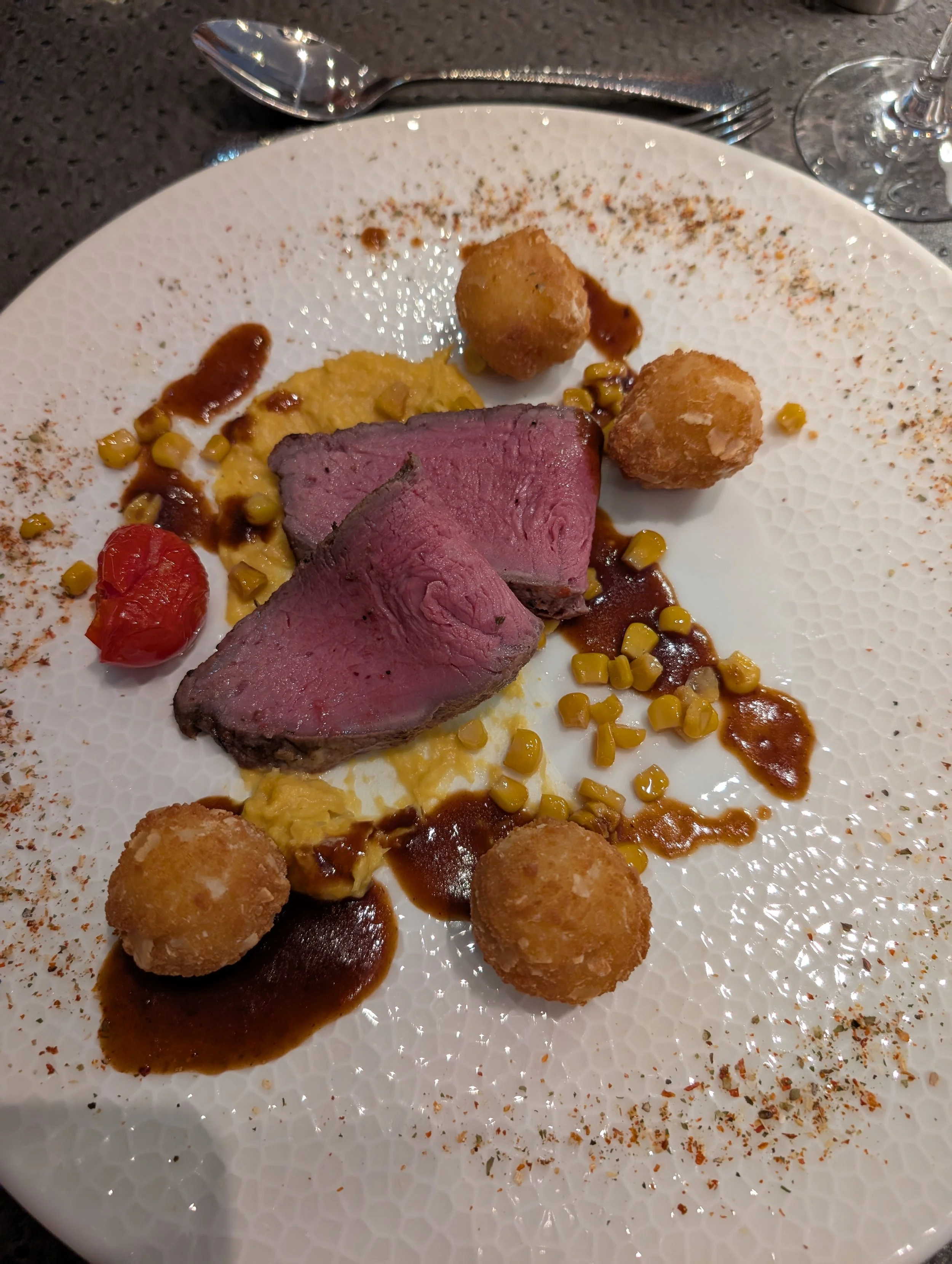 Auf dem Teller befinden sich zwei Scheiben rosa gebratenes Steak, kleine goldbraune Kroketts, eine Kirschtomate, eine Portion cremiges Püree, Erbsen, braune Soße und eine geschmierte Masse, möglicherweise Polenta, alles kunstvoll arrangiert.