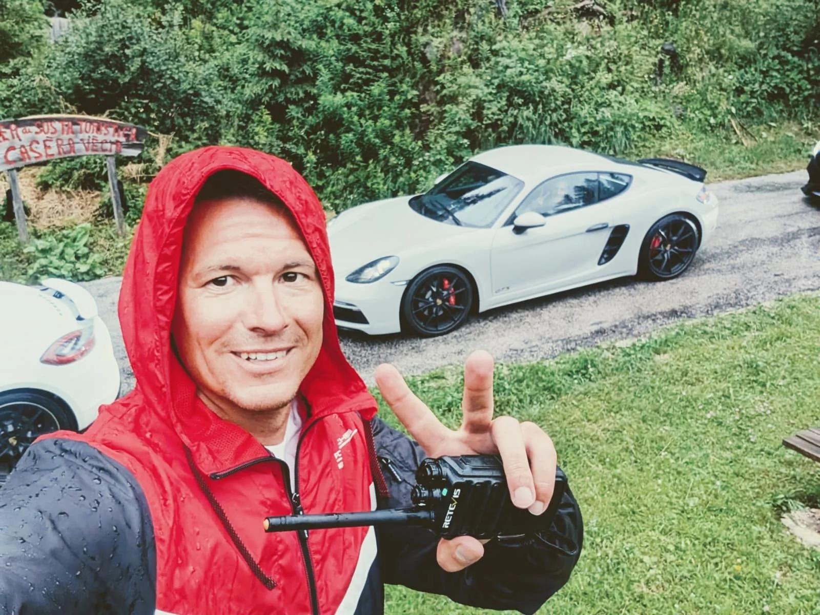 Ein Mann in roter Regenjacke macht ein Selfie vor einem weißen Sportwagen, im Hintergrund viele Bäume und ein Schild mit der Aufschrift 'Casera Velo'