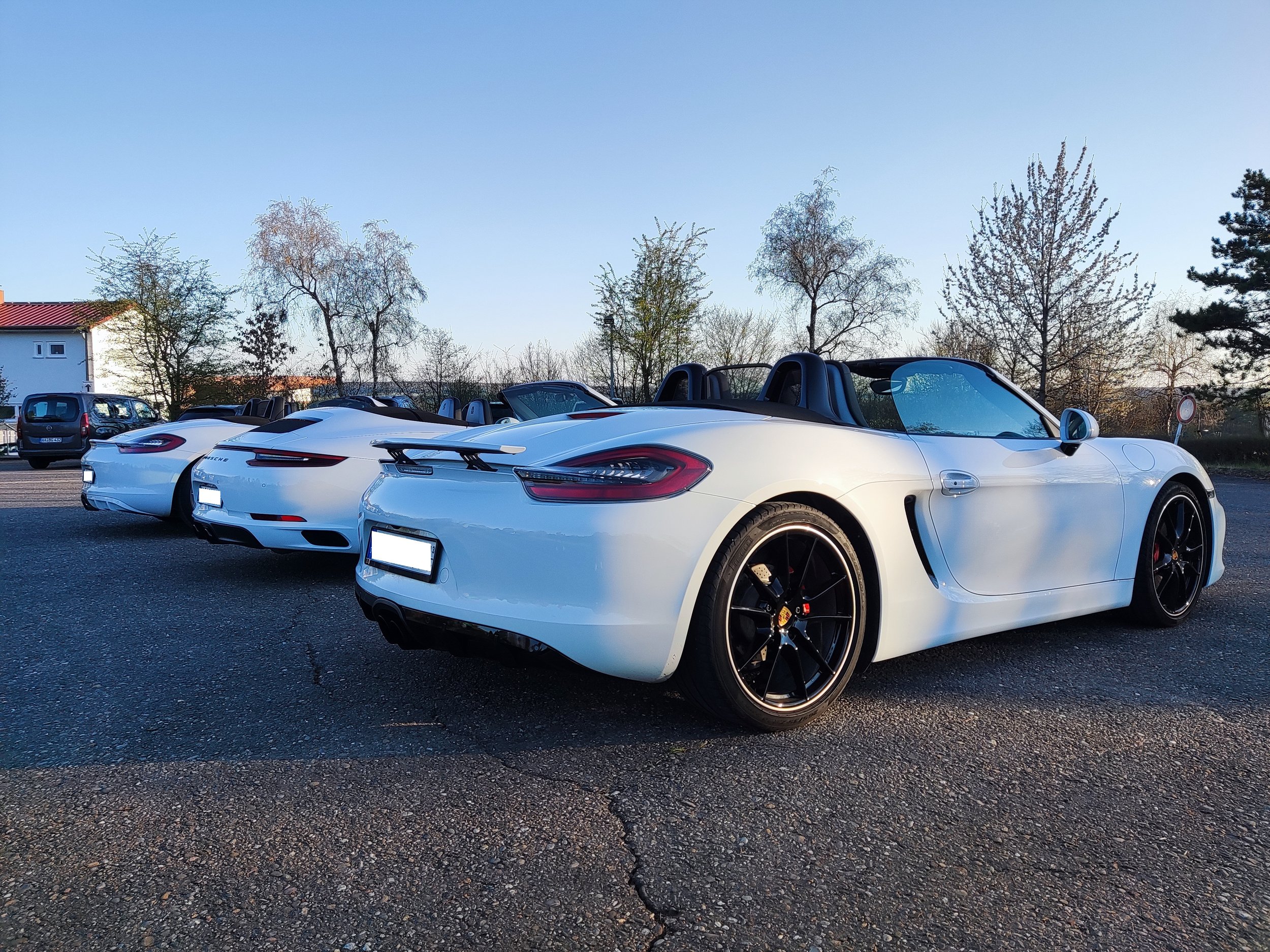 Drei weiße Porsche-Sportwagen mit offenem Verdeck, parkiert auf einer asphaltierenden Fläche mit Bäumen im Hintergrund, bei Sonnenuntergang.