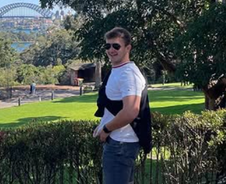 Junger Mann mit Sonnenbrille steht in einem Park, hält eine schwarze Jacke und trägt ein weißes T-Shirt und Jeans.