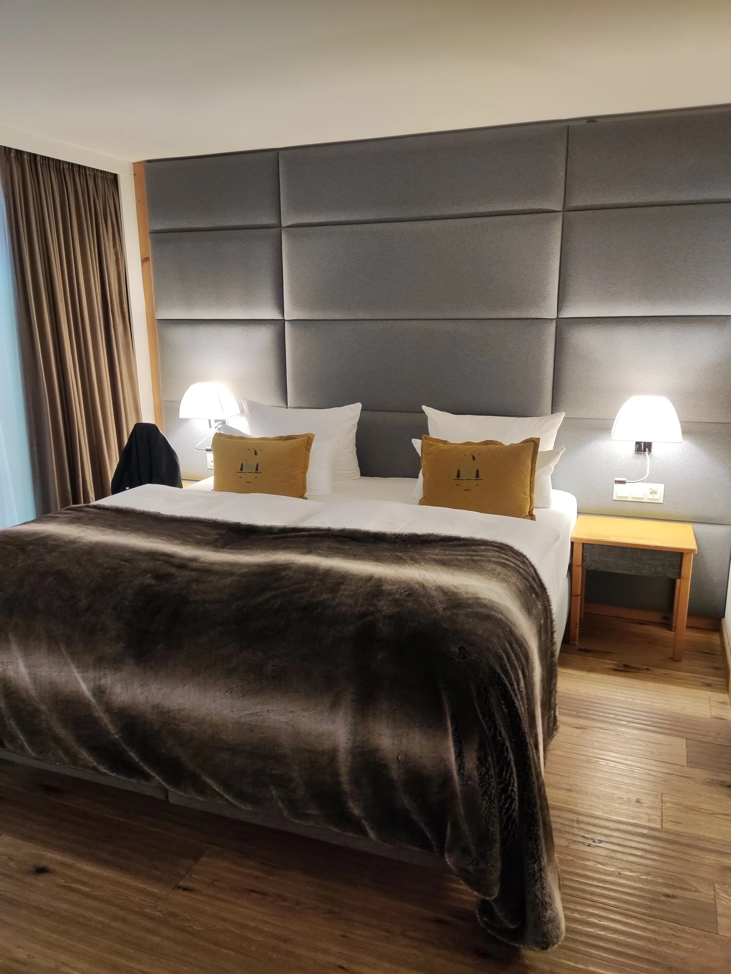 Ein modernes Hotelzimmer mit einem großen Bett, grauem gepolstertem Kopfteil, weißen Bettwäsche, knallgelben Kissen mit einem Waldmotiv, seitlichen Nachttischen aus Holz, und zwei Wandlampen.