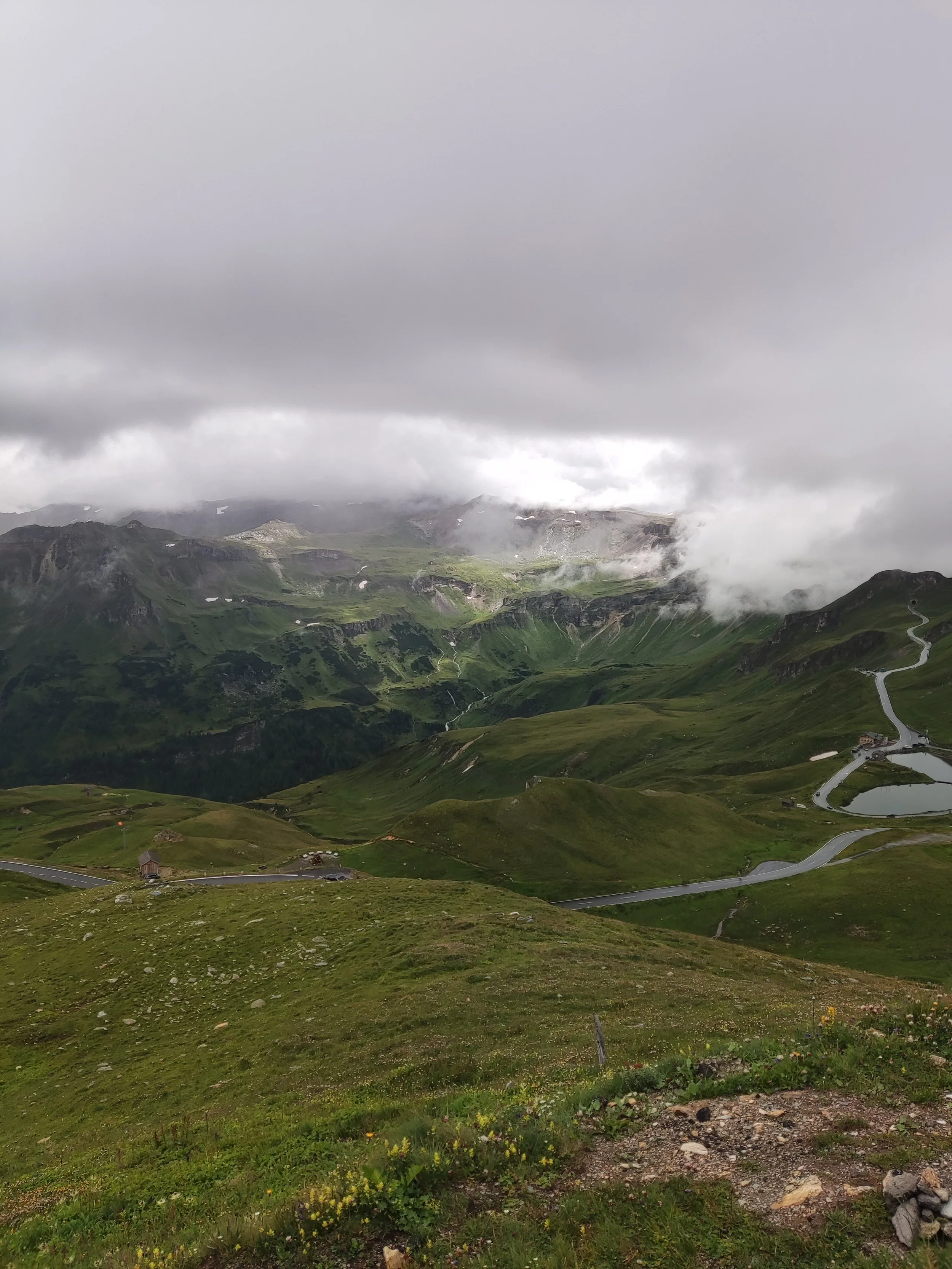Berglandschaft mit grünen Hügeln, Wiesen, einem kleinen See, kurvigen Straßen und bewölktem Himmel.