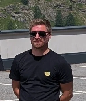 Ein Mann mit dunklem T-Shirt und Sonnenbrille steht am Straßenrand vor einer Mauer und einem bewaldeten Hang.