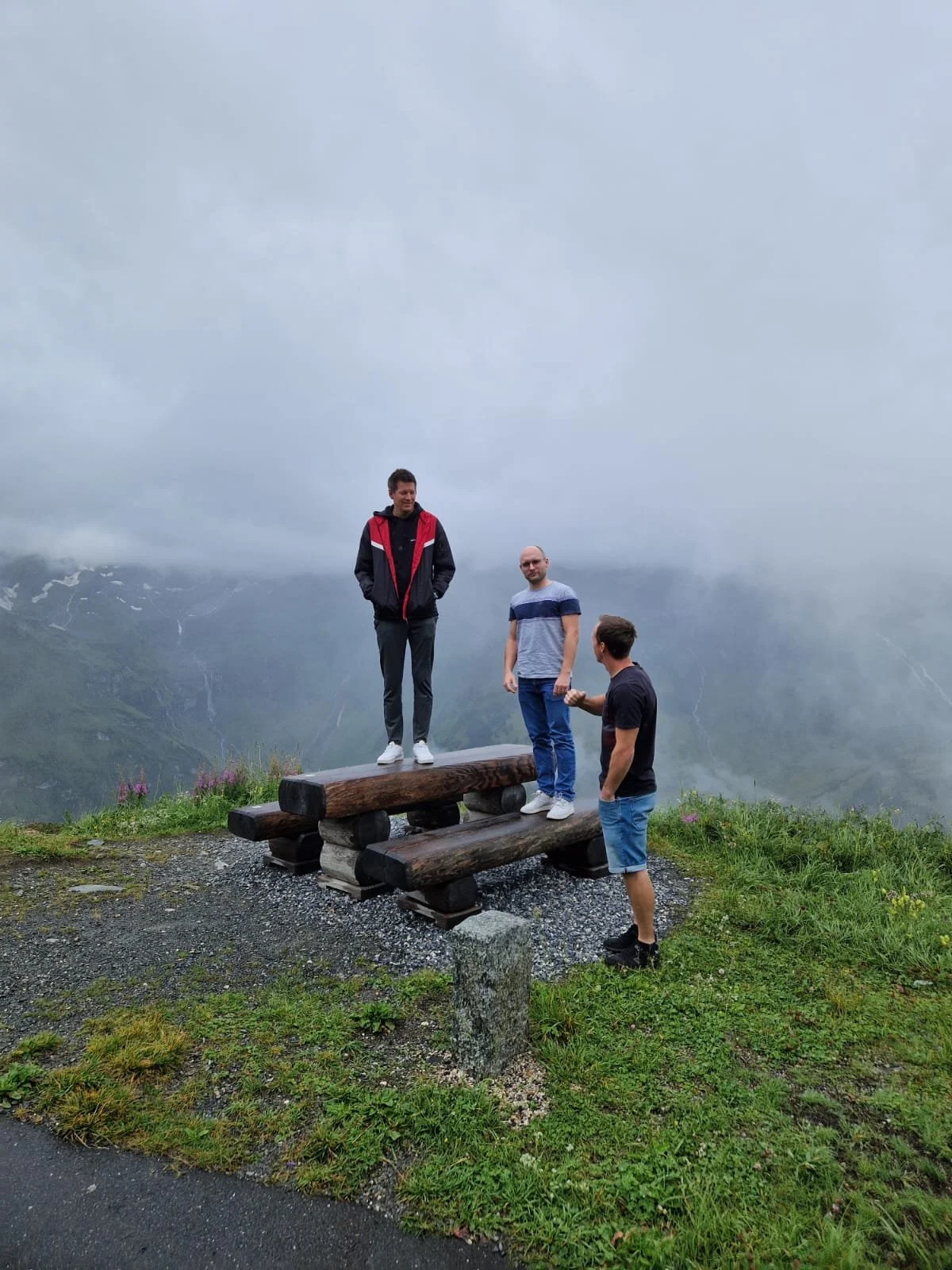 Drei junge Männer stehen auf einem Holzpodest in einer bergigen, nebligen Landschaft.